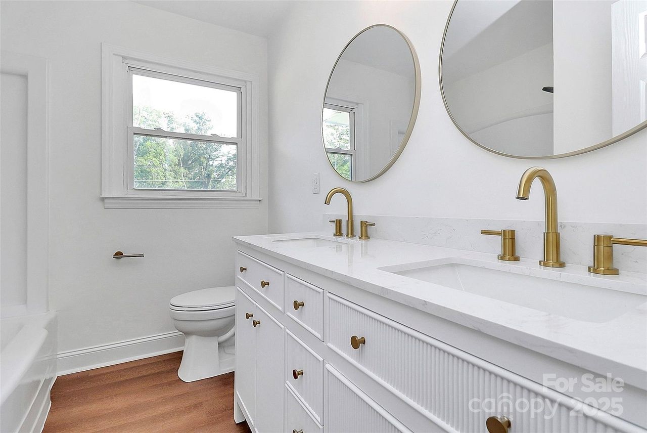 Bright white bathroom with wooden floor, double-sink vanity featuring gold faucets and hardware, oval mirrors, a window above toilet, and a bathtub visible on side.