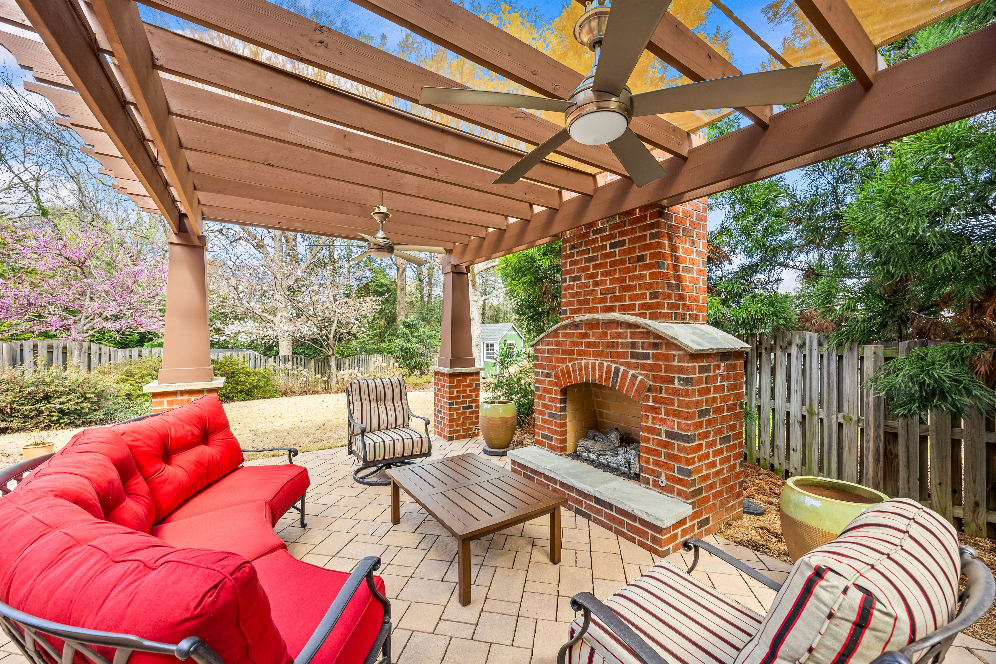 Backyard covered patio with brick outdoor fireplace, wooden pergola, ceiling fans, red cushioned sofa and striped chairs around a brown wooden coffee table; fenced garden with trees in bloom.