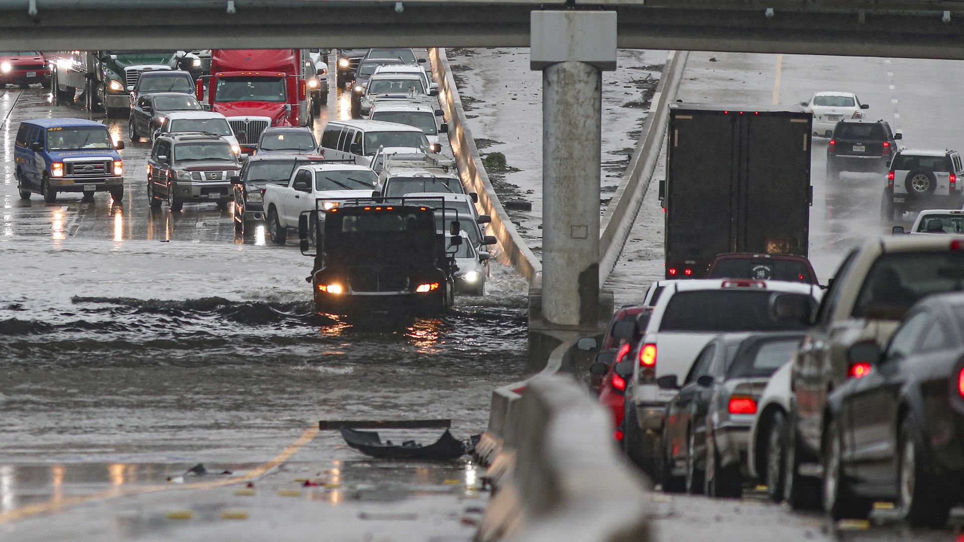 In this image, a truck drives under an overpass with a trail of other cars behind it.