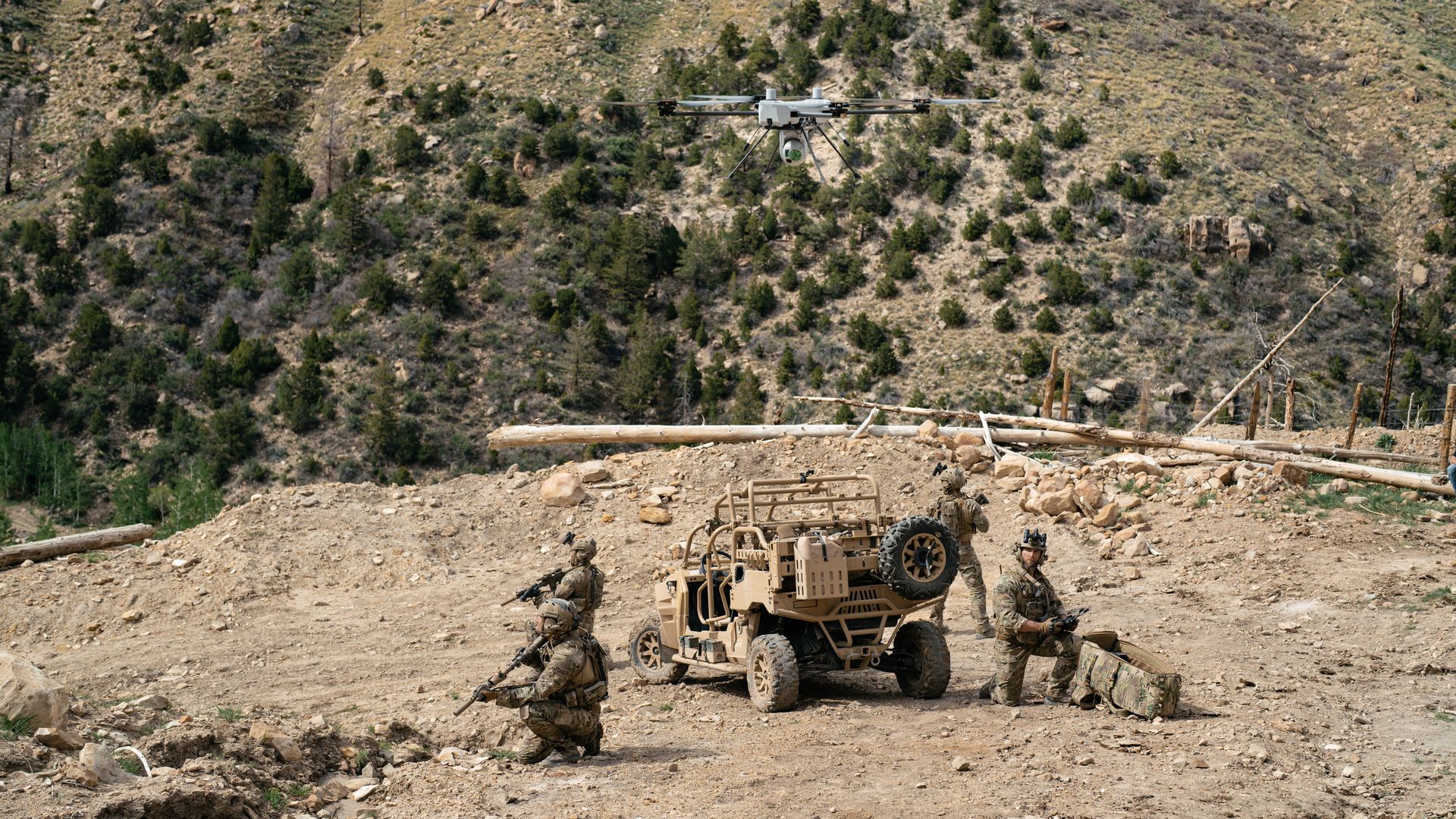 A drone hovers over troops, who are crouched by an off-road vehicle. They are in the mountains.