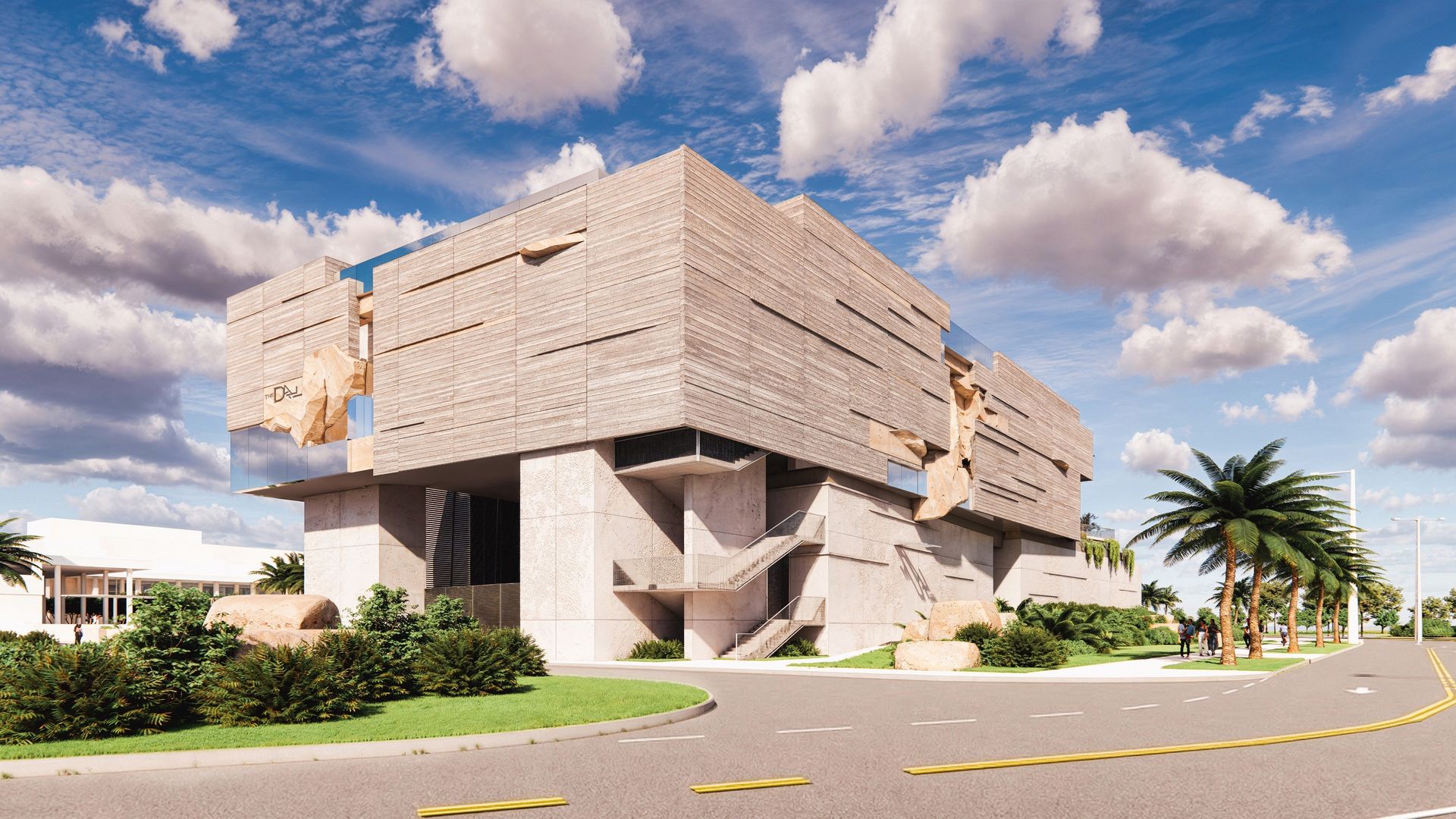 Modern beige concrete building with layered textured upper section and large boulders attached, surrounded by green plants and palm trees under a blue sky with white clouds.