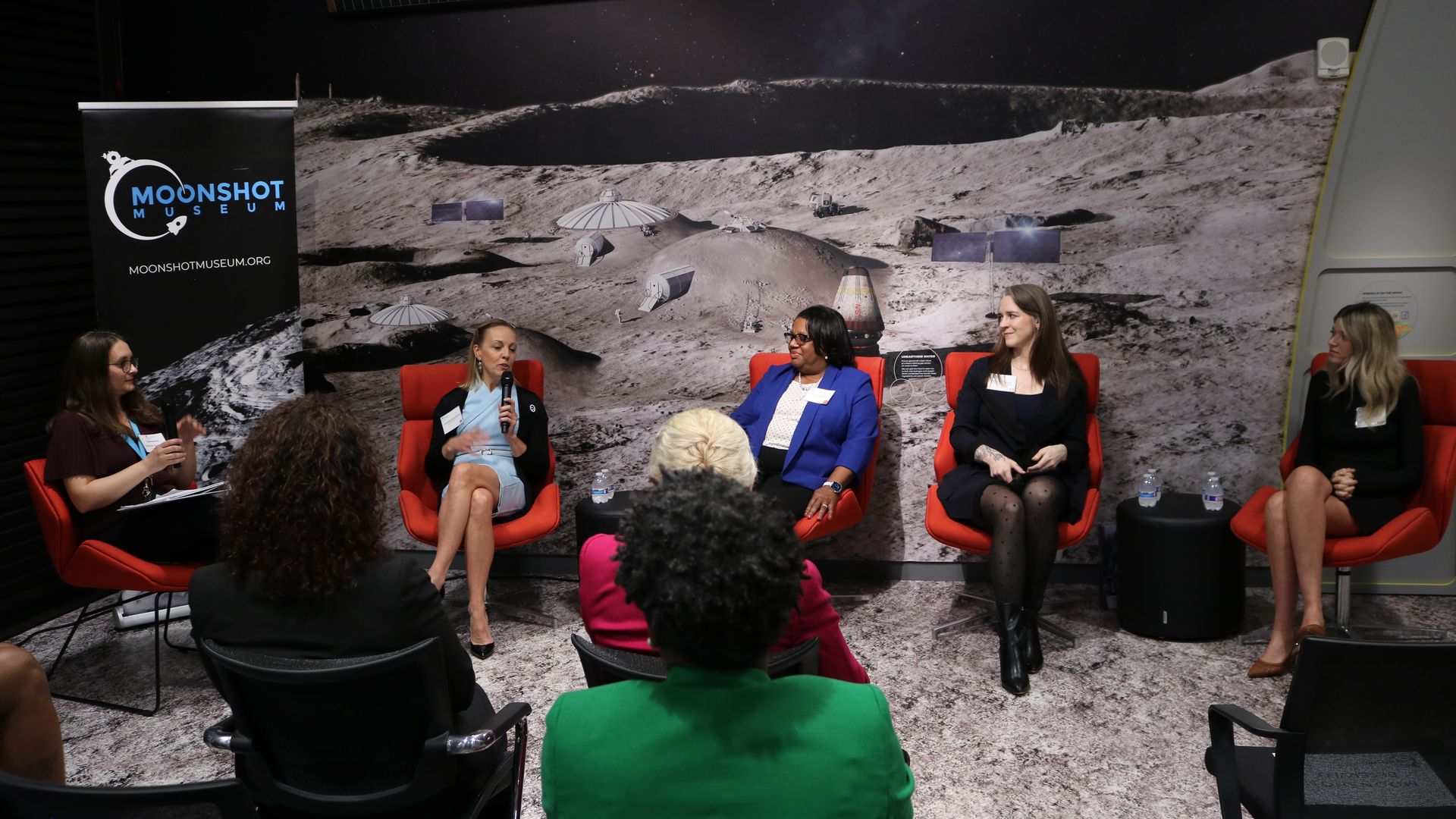 Panel of five women seated in red chairs on a Moonshot Museum stage, with one speaking into a microphone; a lunar surface backdrop and Moonshot banner behind them.