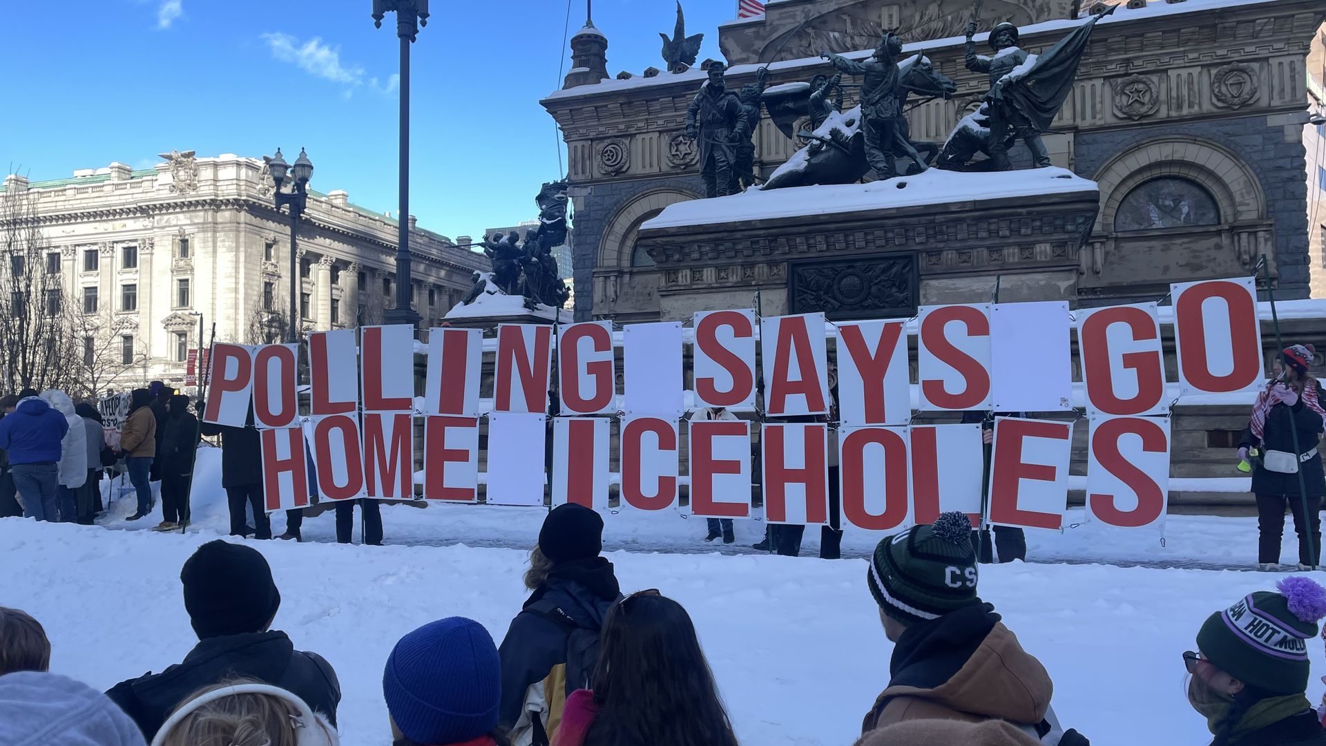 People standing in snow holding large red and white signs reading "POLLING SAYS GO HOME LICEHOLES" in front of a snowy statue and historic building on a clear day.