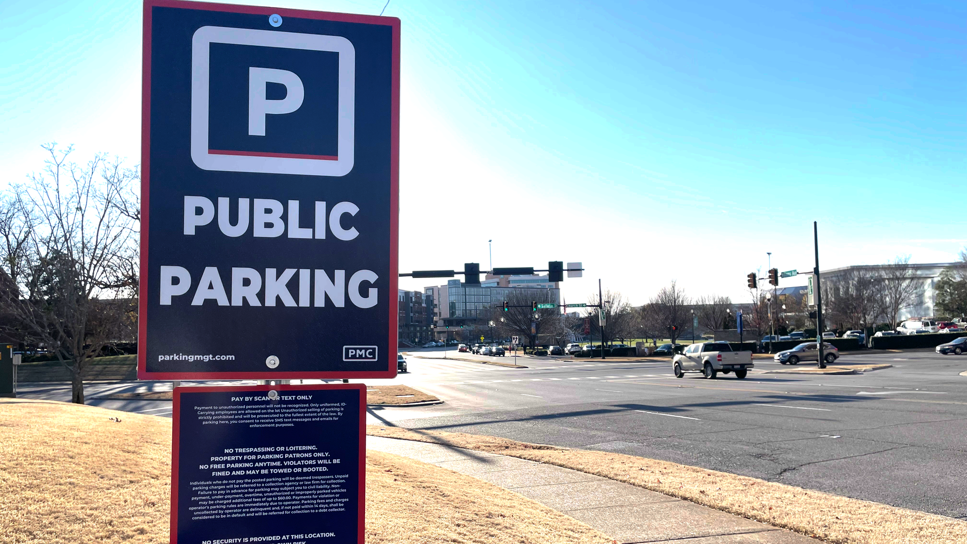 Blue and white public parking sign with rules on a grassy area near a busy intersection with cars and buildings under a clear blue sky.