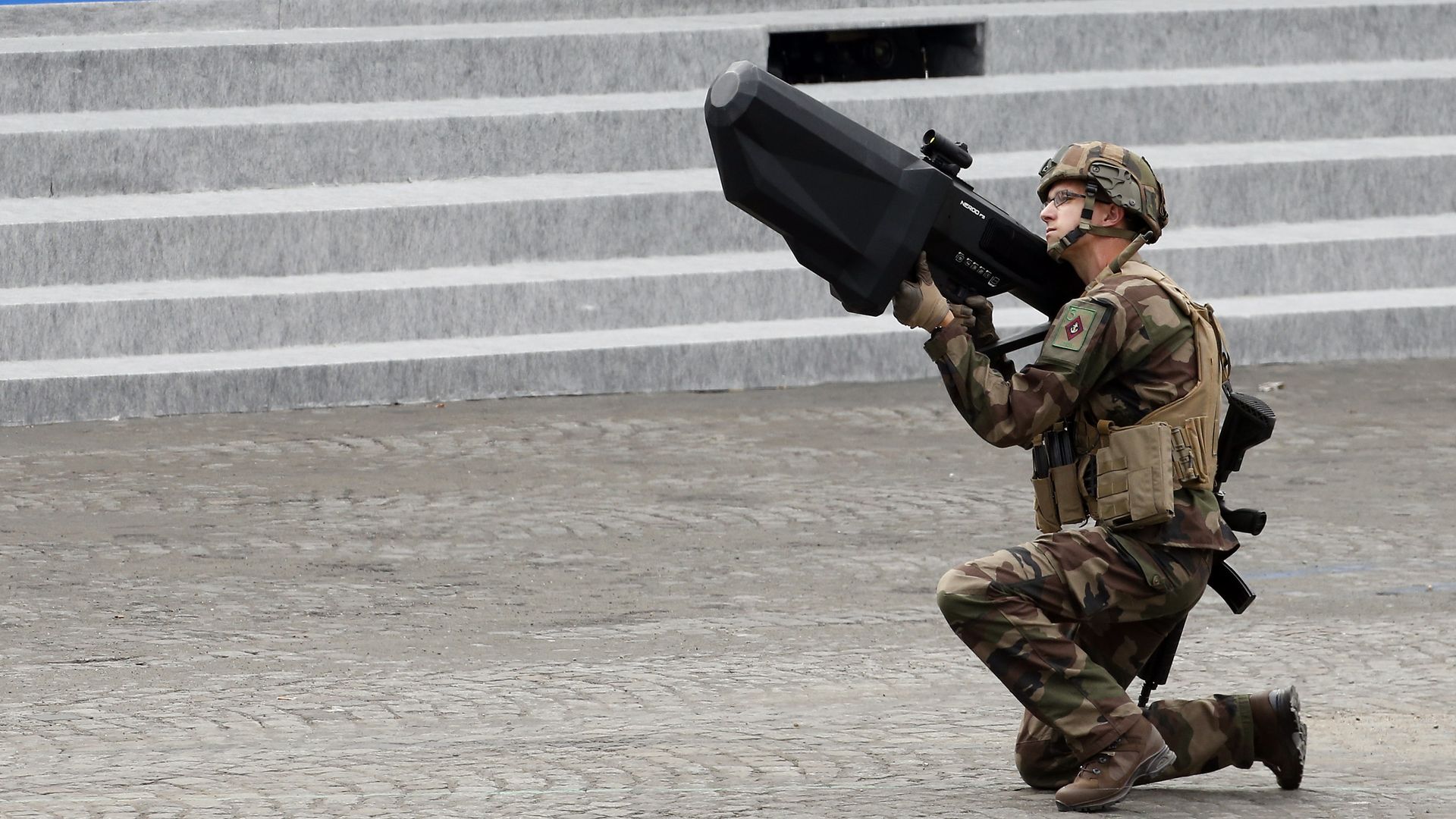 A crouching man in fatigues points a rifle-like object toward the sky