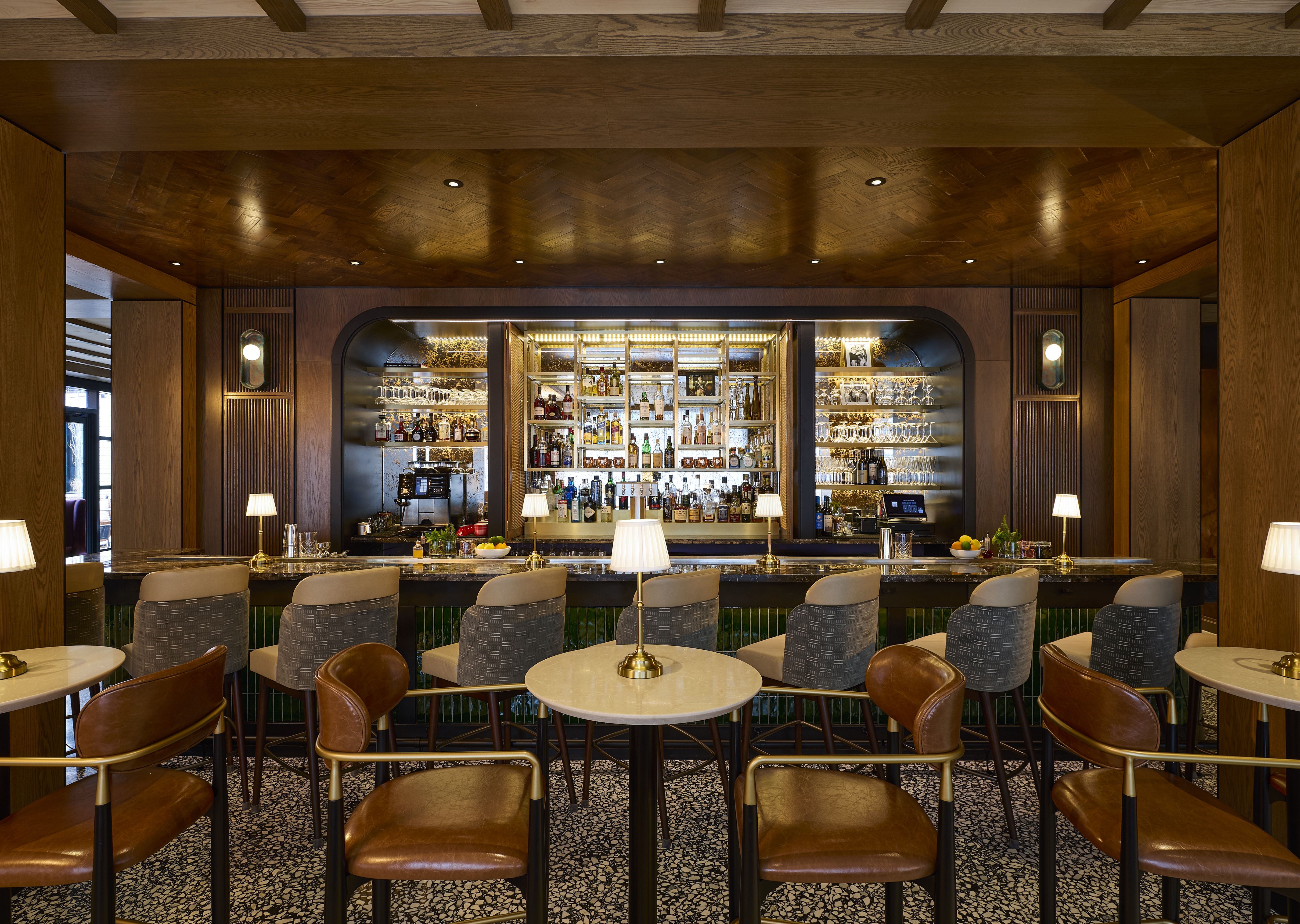 Elegant bar interior with wood-paneled ceiling and walls, marble tables, leather and fabric chairs, and shelves of bottles and glasses illuminated by warm lighting.