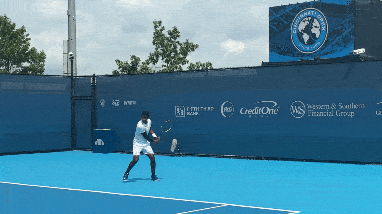 Vignesh Gogineni, wearing all whites and black sneakers, warms up from the baseline on a court at the Lindner Family Tennis Center in Cincinnati, Ohio.