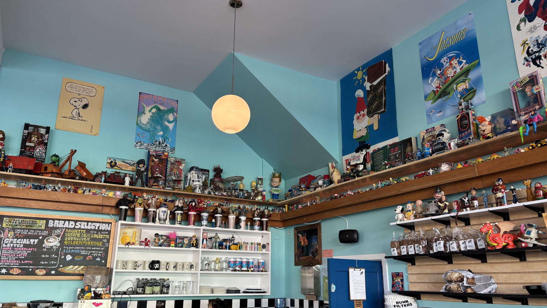 Teal-walled toy shop with a round hanging lamp, retro posters like "The Jetsons" and Snoopy, shelves of vintage figures and cars, plus a chalkboard menu and pastry counter.
