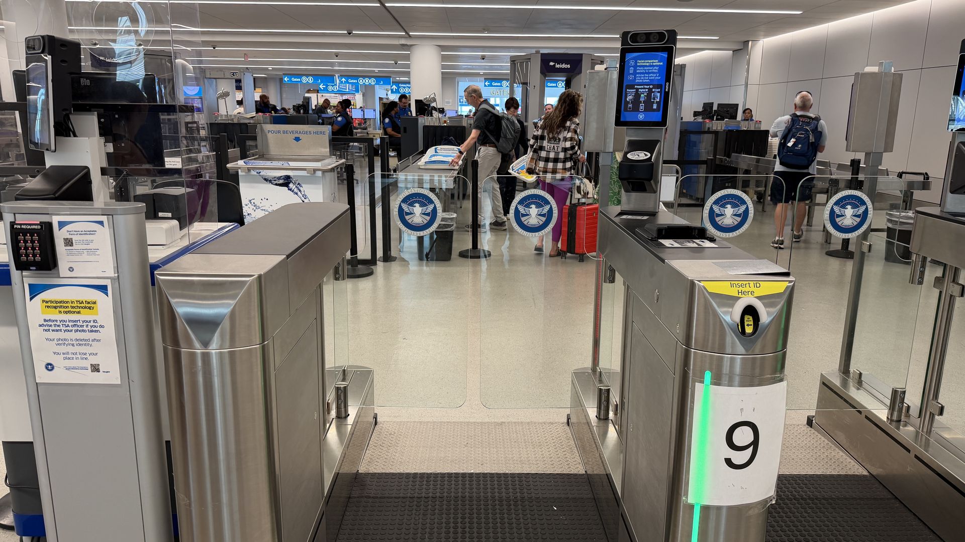 Busy airport security checkpoint with metal gates, glass barriers, and TSA logos. Travelers with rolling luggage wait in line beneath blue signs and a tall digital information screen.