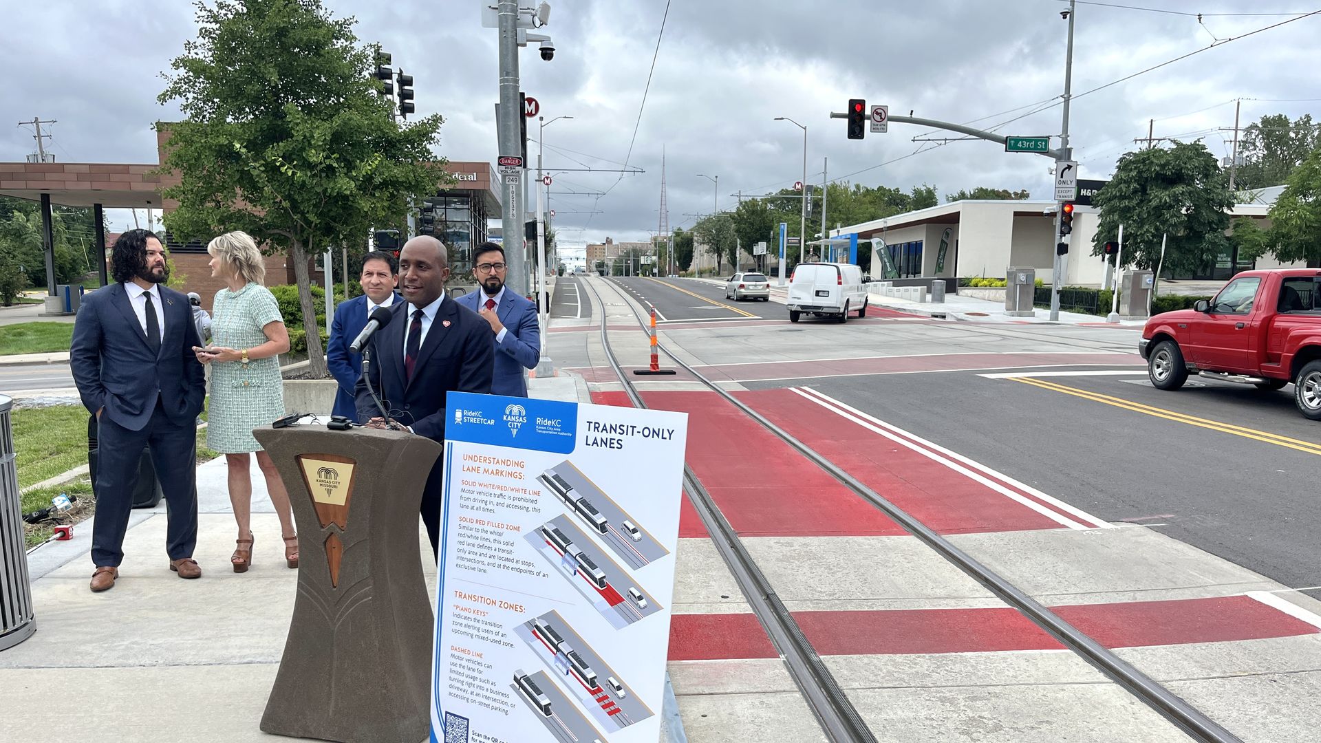Man in suit speaking at a podium with a transit lanes explanation sign, four people standing behind, streetcar tracks and red transit-only lanes visible on an urban street with cloudy sky.