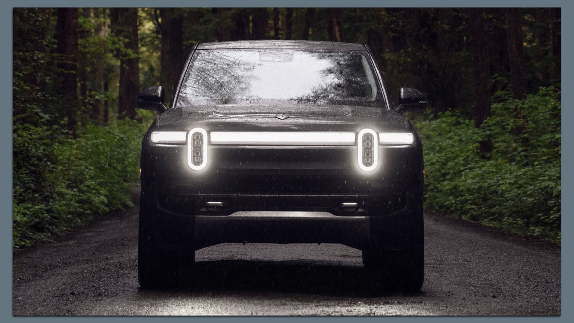 Front view of a dark Rivian electric SUV with vertical LED headlights and a glowing horizontal light bar, parked on a wet forest road. Rain beads on the windshield; dense trees frame the scene.