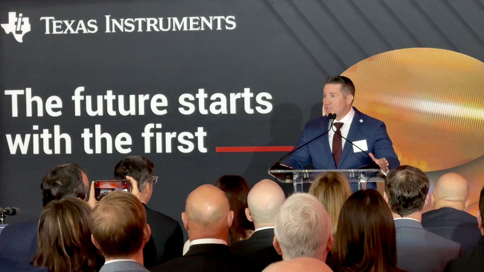 Speaker in a blue suit at a podium on stage; a large screen shows the Texas Instruments logo and the words "The future starts" while an audience looks on.