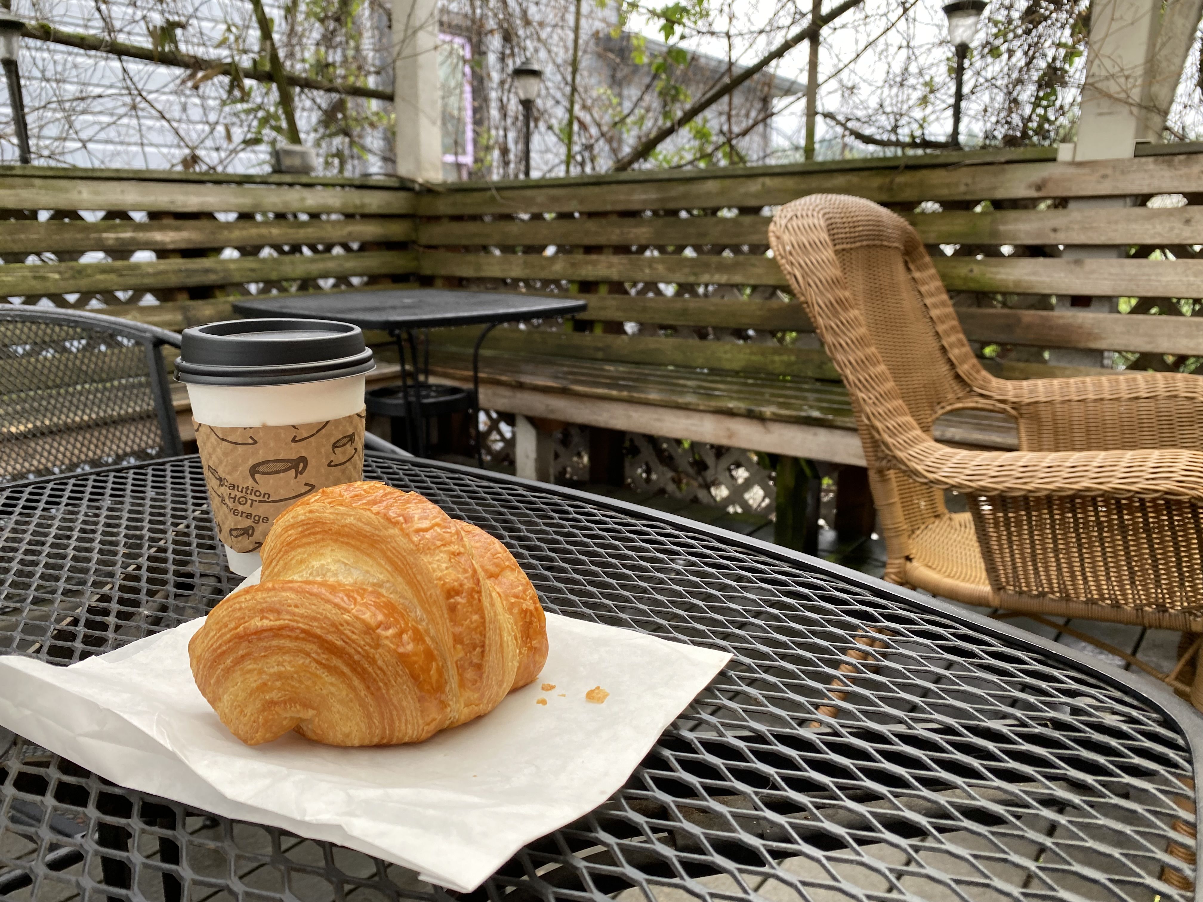 A croissant and a cup of coffee in a to go cup sitting on a metal outdoor cafe table with wooden wall and hanging plants in background.