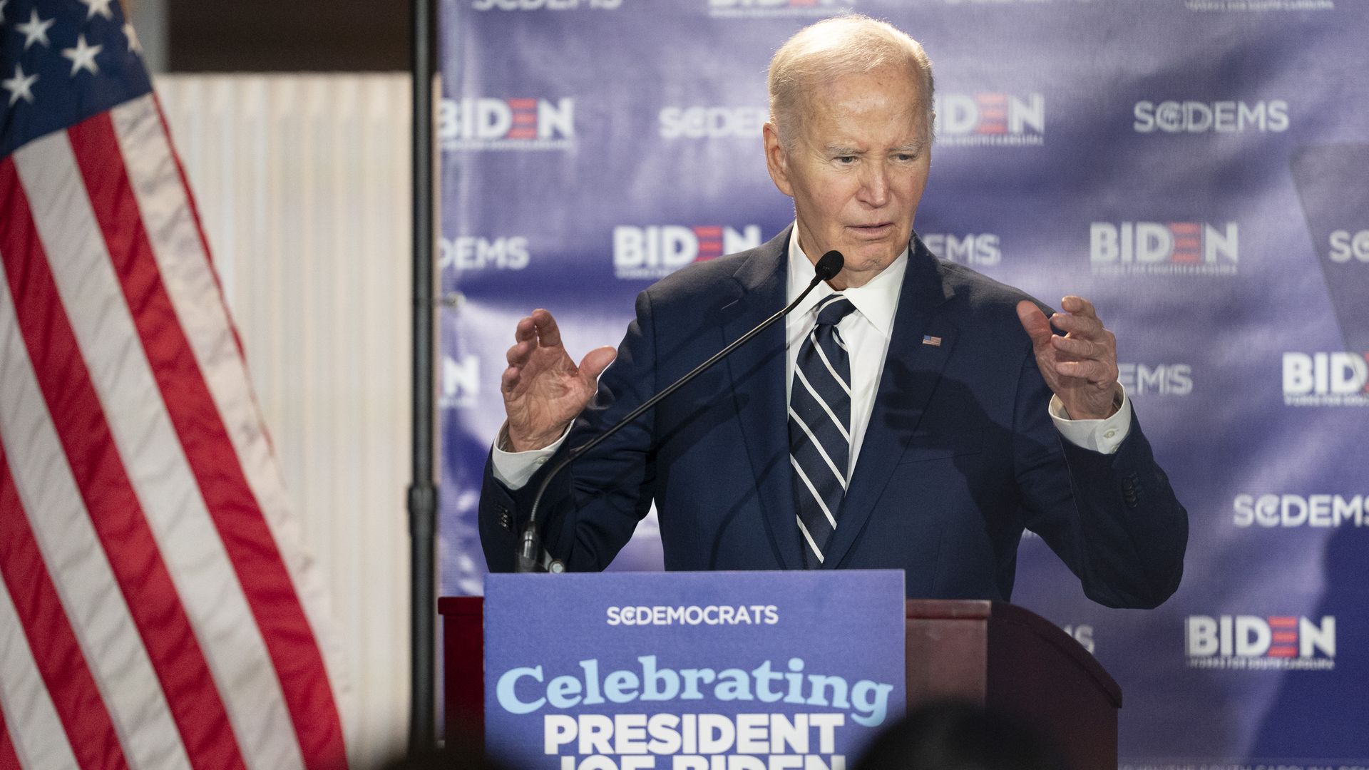 Joe Biden at a podium giving a speech, blue backdrop with BIDEN logos, American flag on the left, wearing a dark suit and striped tie, gesturing with his hands.