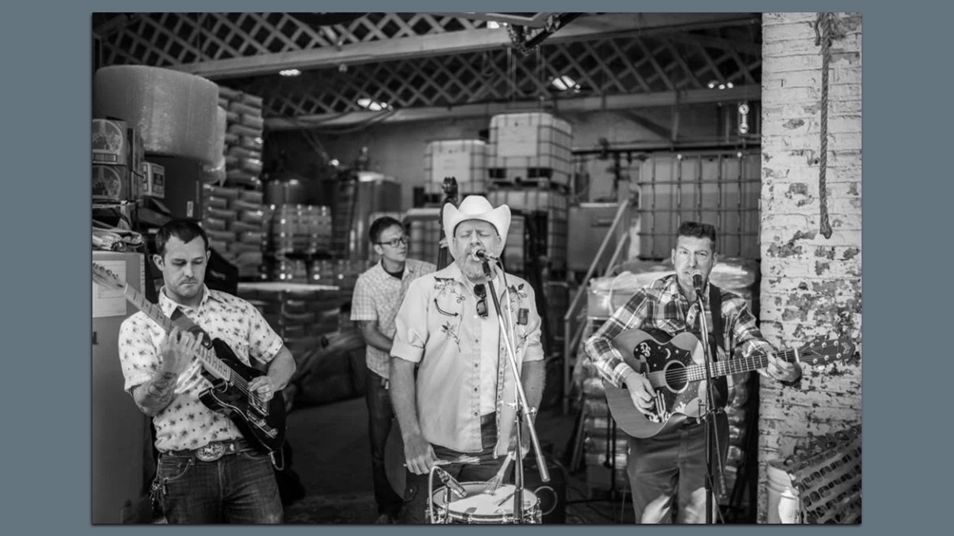 Black-and-white photo of a three-man band performing in a warehouse: left guitarist, center singer in a cowboy hat at a mic, right guitarist with an acoustic guitar; pallets and a brick wall behind.
