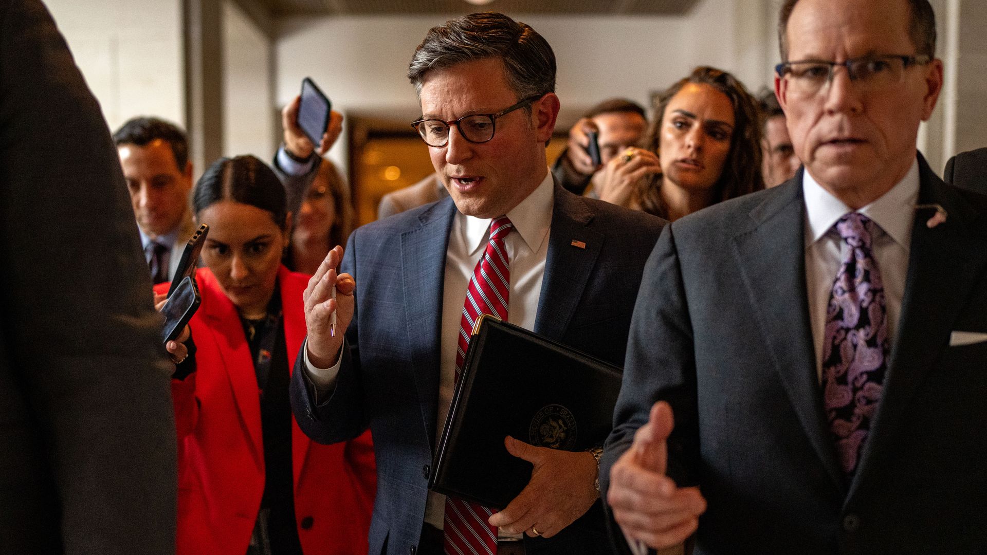Speaker Mike Johnson, wearing a gray suit, walking with a gaggle of reporters through a white hallway.