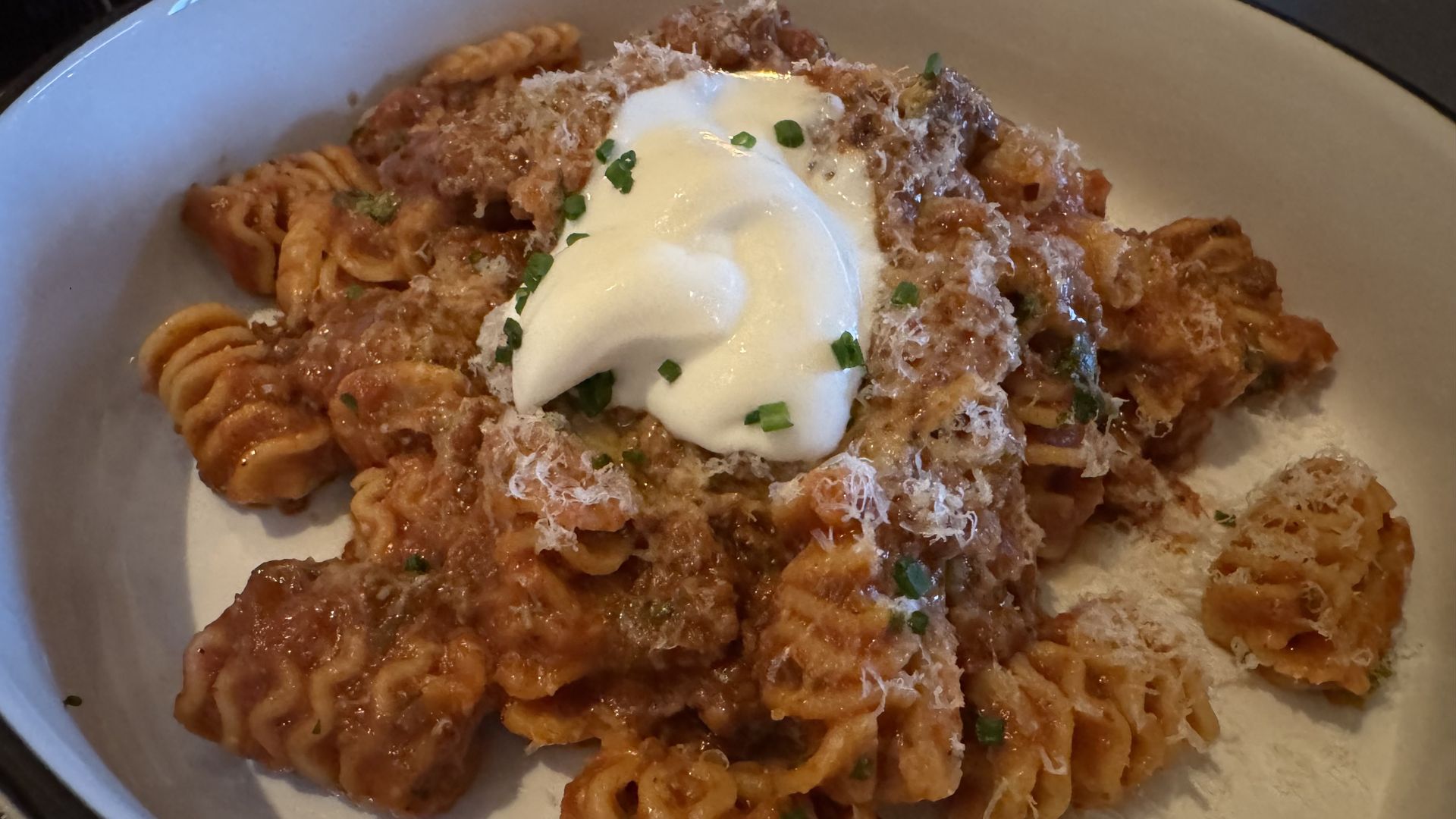 A bowl of rotini pasta in red sauce topped with grated cheese, herbs, and a dollop of creamy white topping.