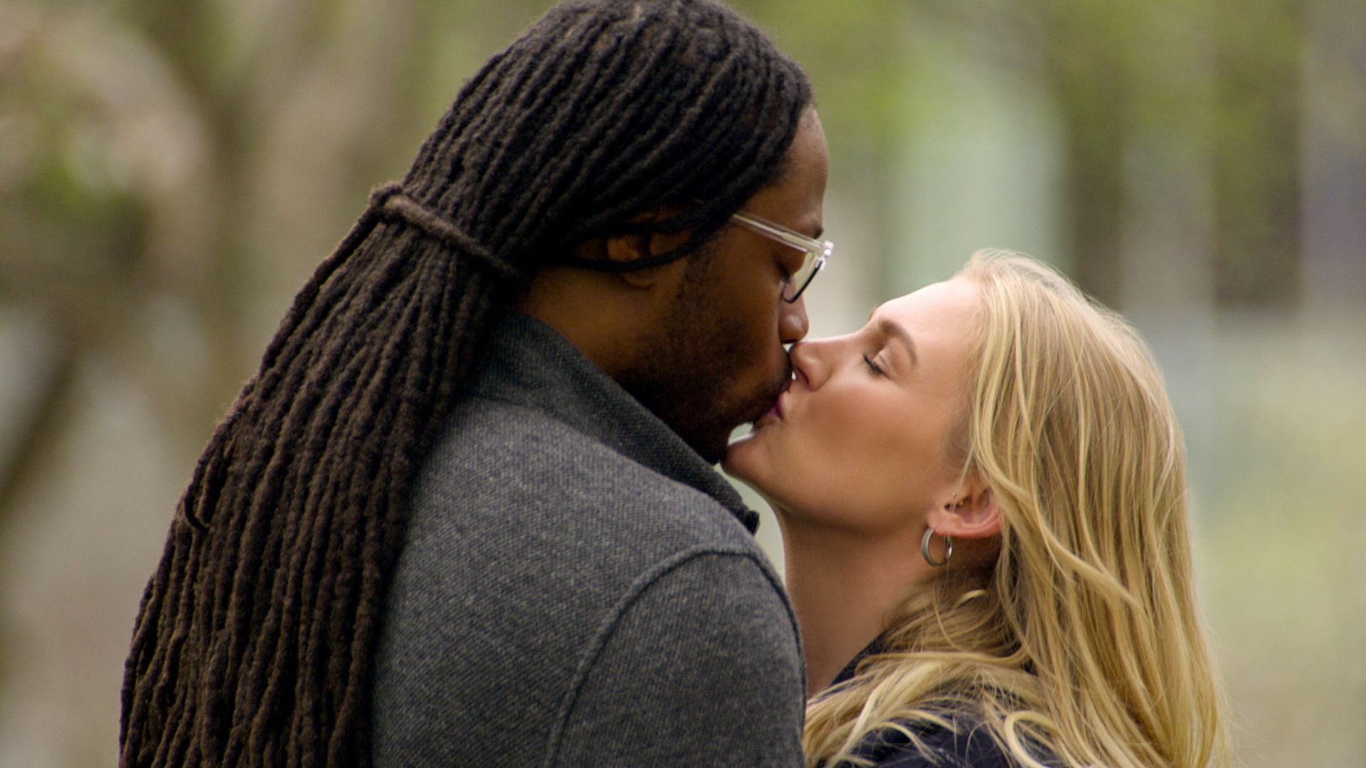 A Black man with long dreadlocks and glasses kissing a blonde white woman outdoors, both eyes closed, surrounded by blurred green trees.