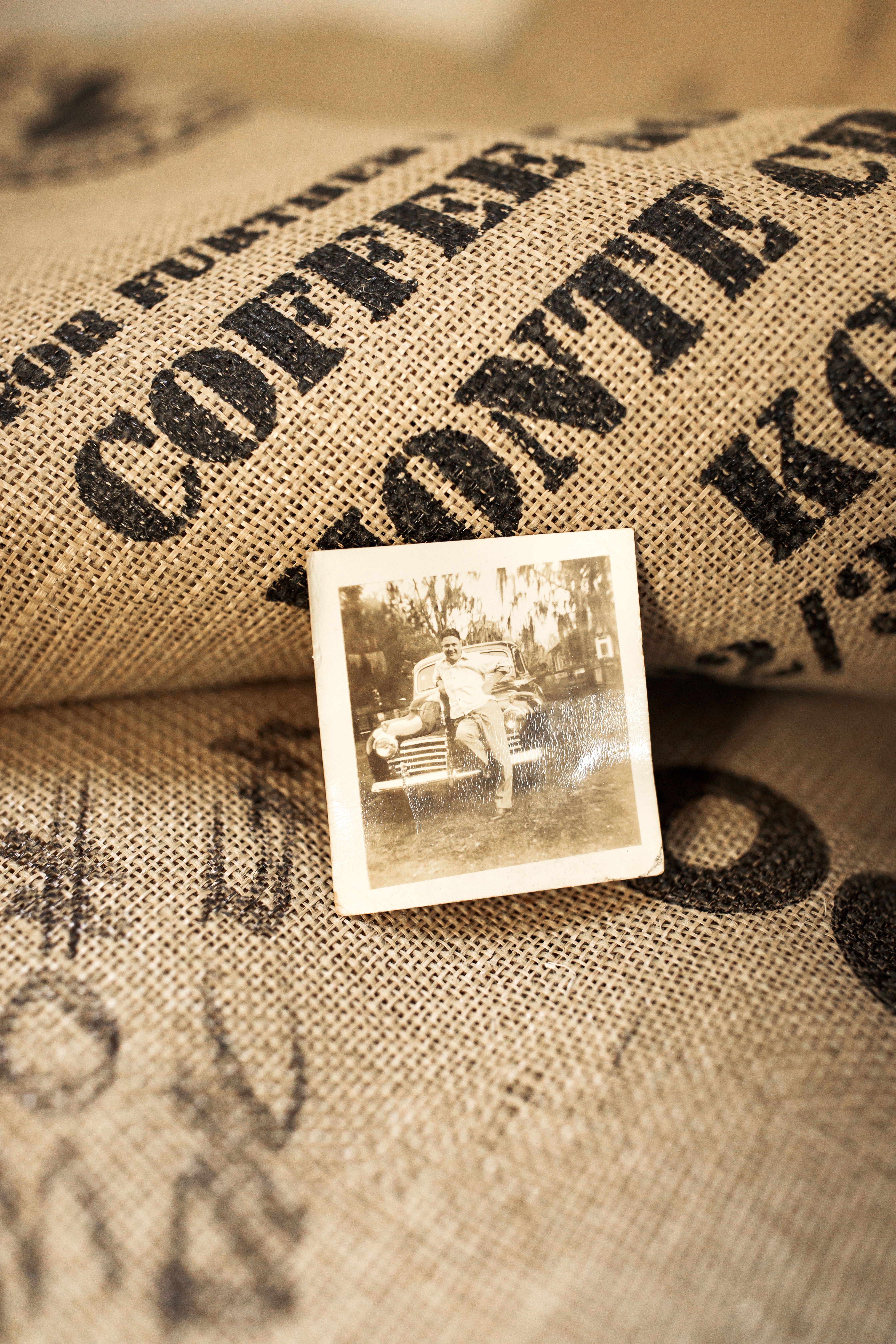 A black and white image of a man leaning against a car sits on top of burlap coffee sacks.