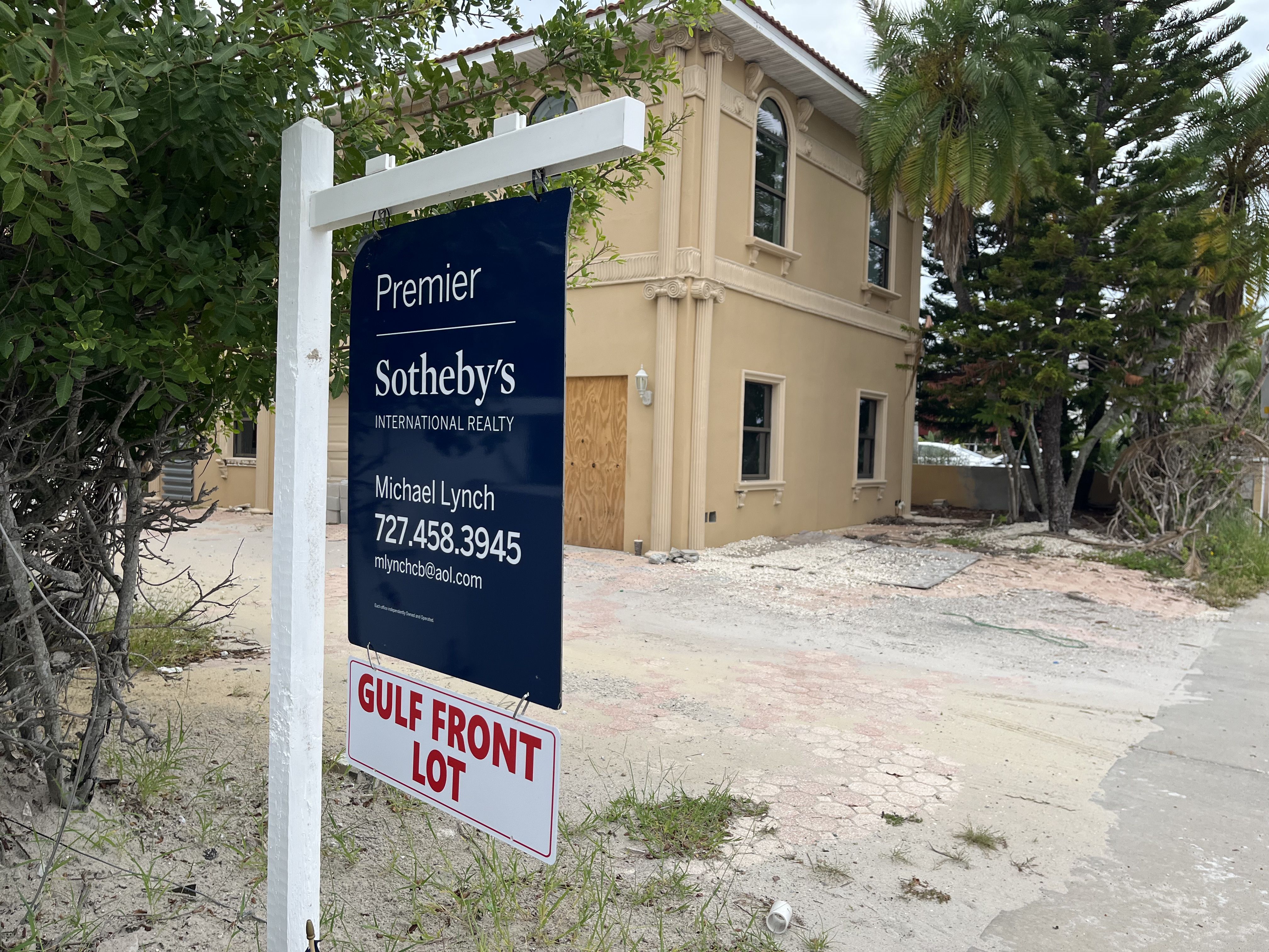 Realty sign for Premier Sotheby's with agent Michael Lynch's contact in front of a beige, boarded-up house with trees around and a sandy, cracked driveway labeled "GULF FRONT LOT".