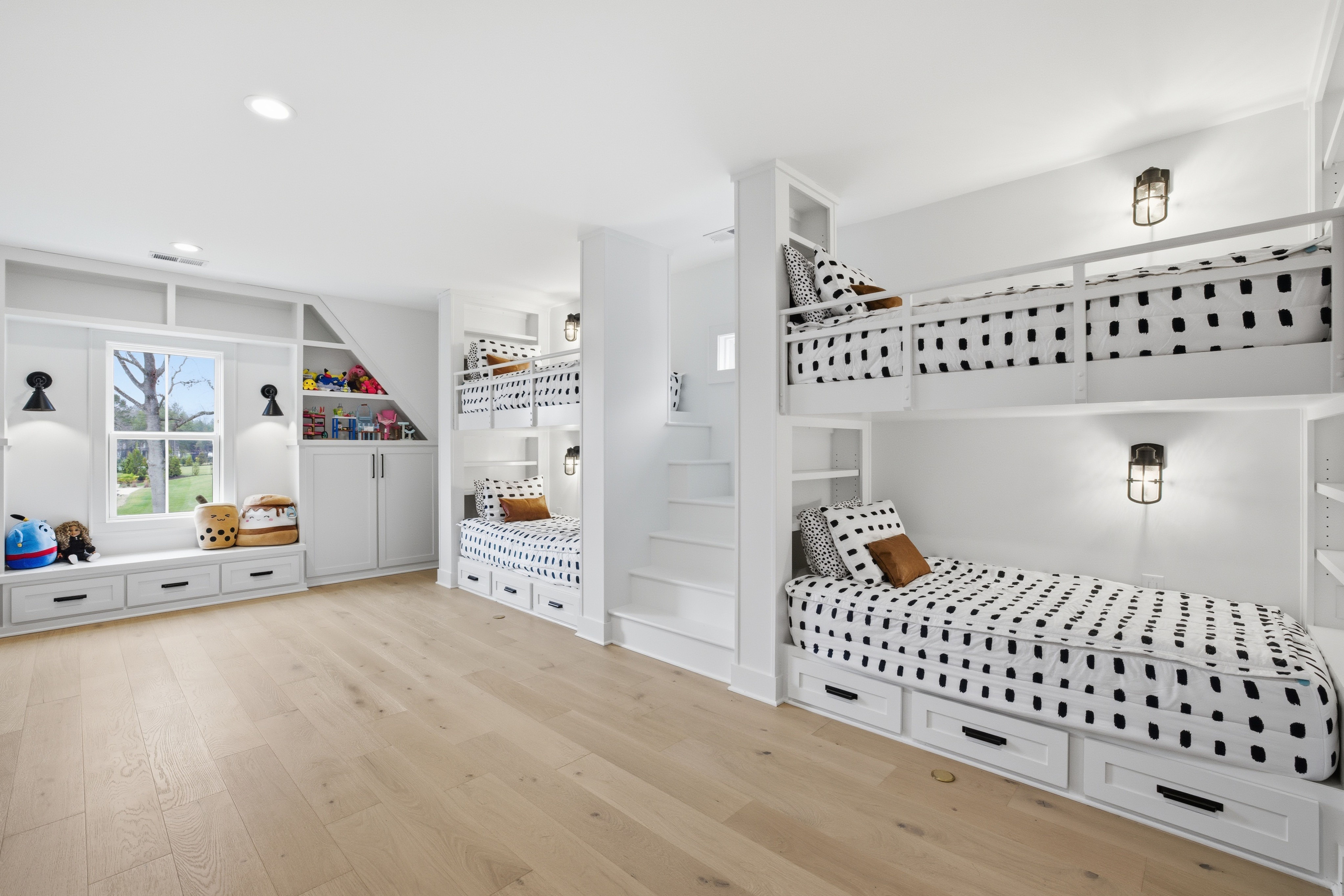 Bright white kids' bedroom with two sets of bunk beds featuring black and white polka dot bedding and brown accent pillows, light wood floor, built-in shelves, and large window with toys.