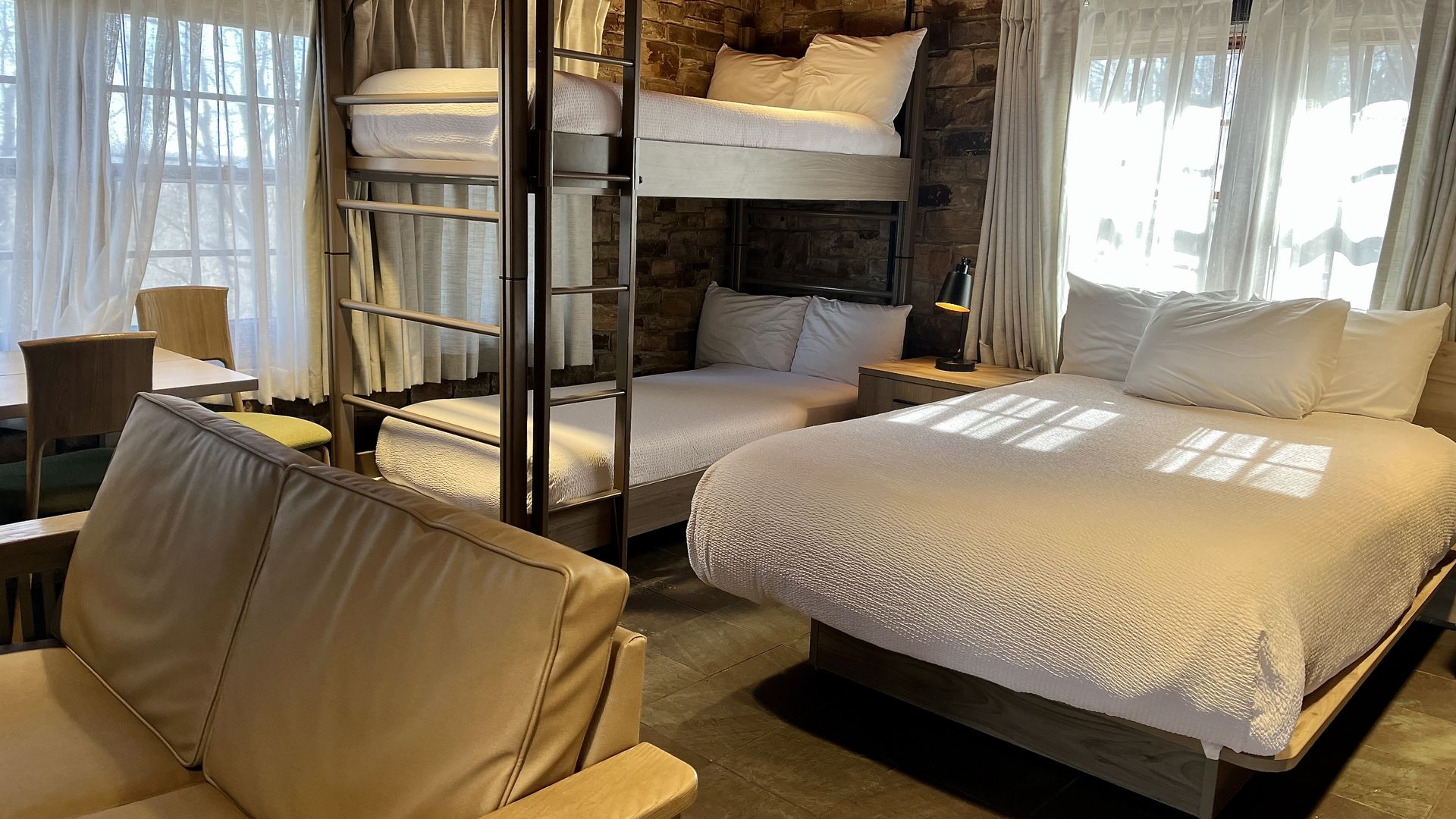 Cozy room with a white bed, bunk beds, tan sofa, wooden ceiling, stone walls, and large windows with sheer white curtains letting in natural light.