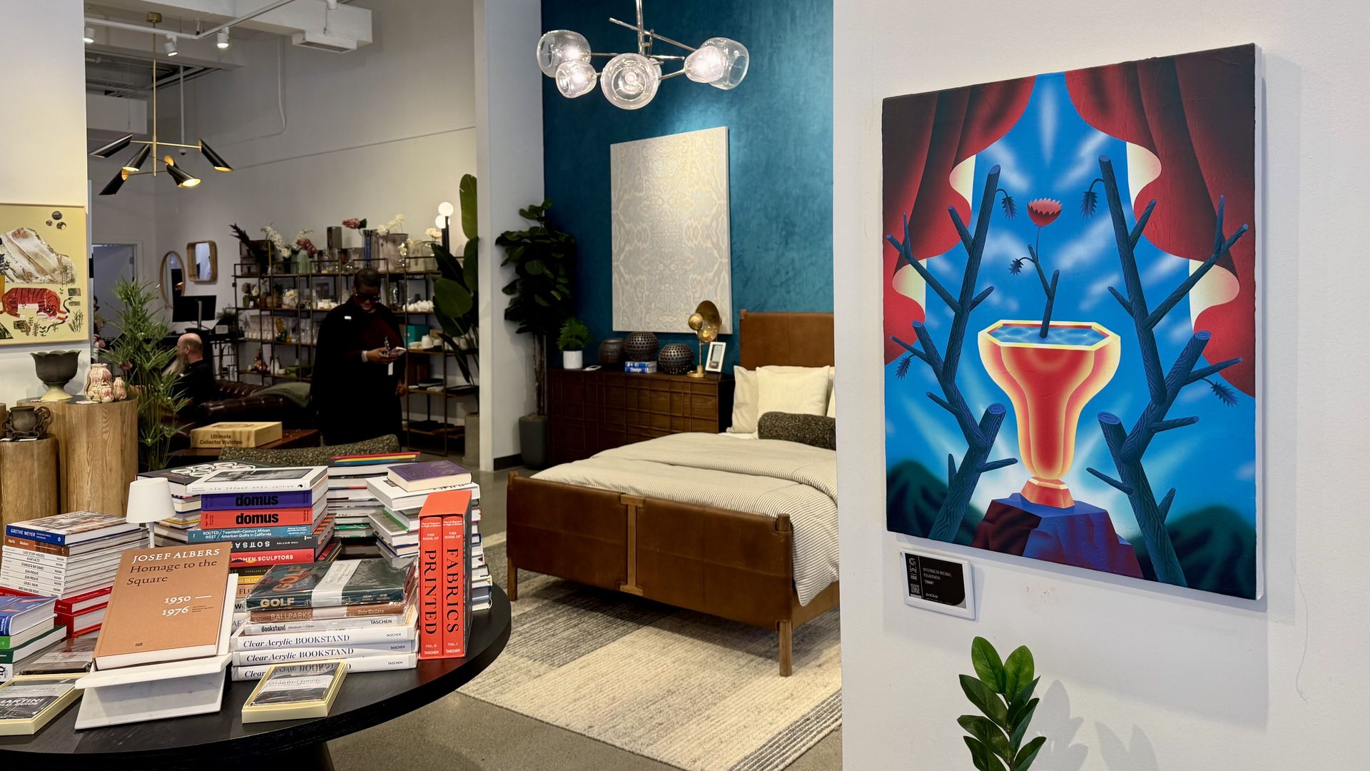 Interior of a modern showroom with a wooden bed against a teal wall, a round table of stacked books in the foreground, a bold painting on the right, and a person browsing shelves behind. 