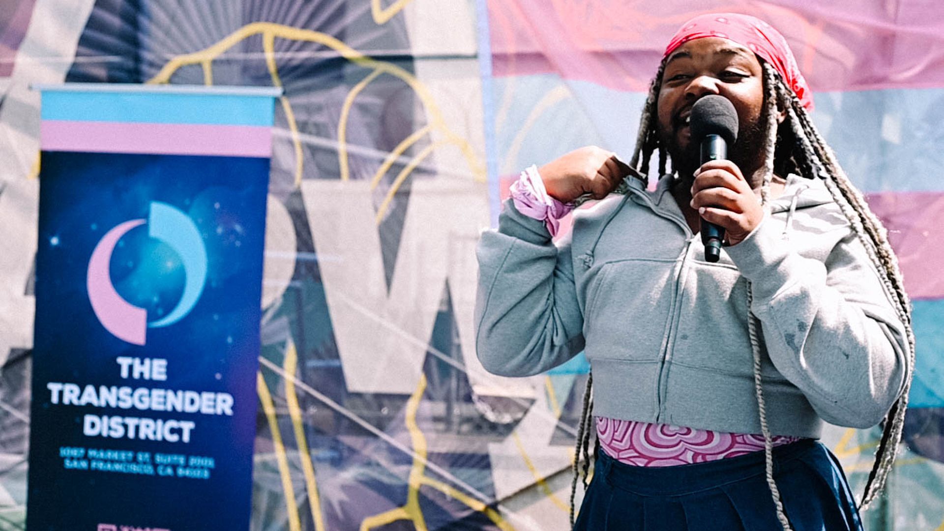 Person with long braided hair and a pink bandana speaking into a microphone, wearing a gray hoodie and navy blue skirt at an event with a banner reading "The Transgender District" in the background.