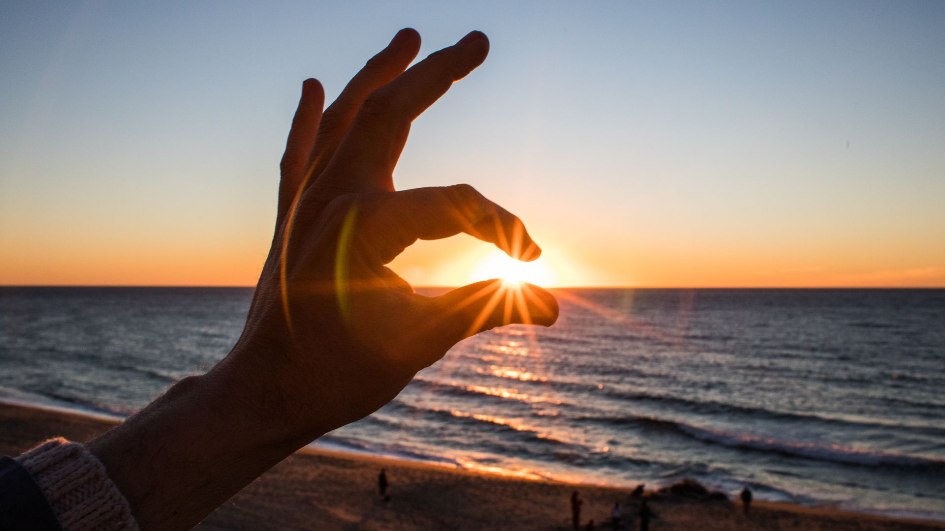 In this image, someone pretends to pinch the sun between two fingers as the sun sets at the beach.