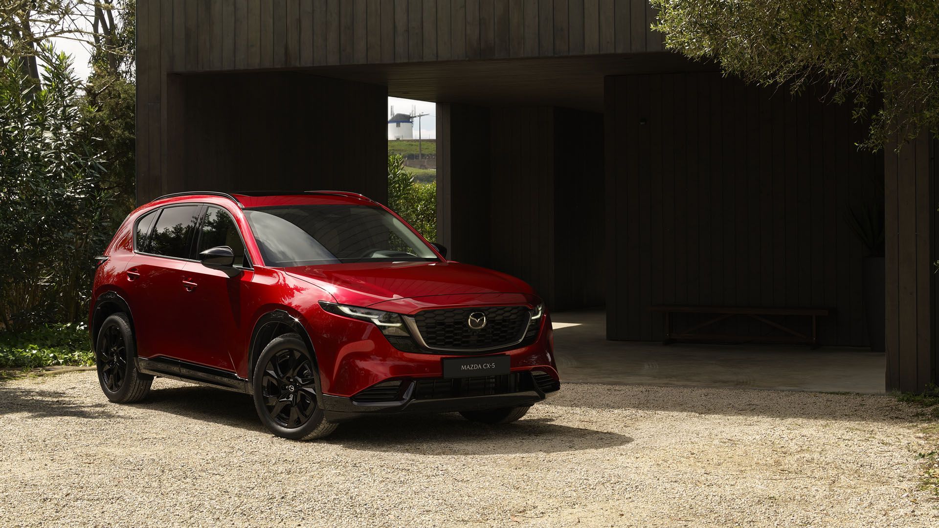 Red Mazda CX-5 SUV parked in a gravel driveway beside a dark wooden building, with green trees on the left and a bright white structure visible through a doorway in the background.