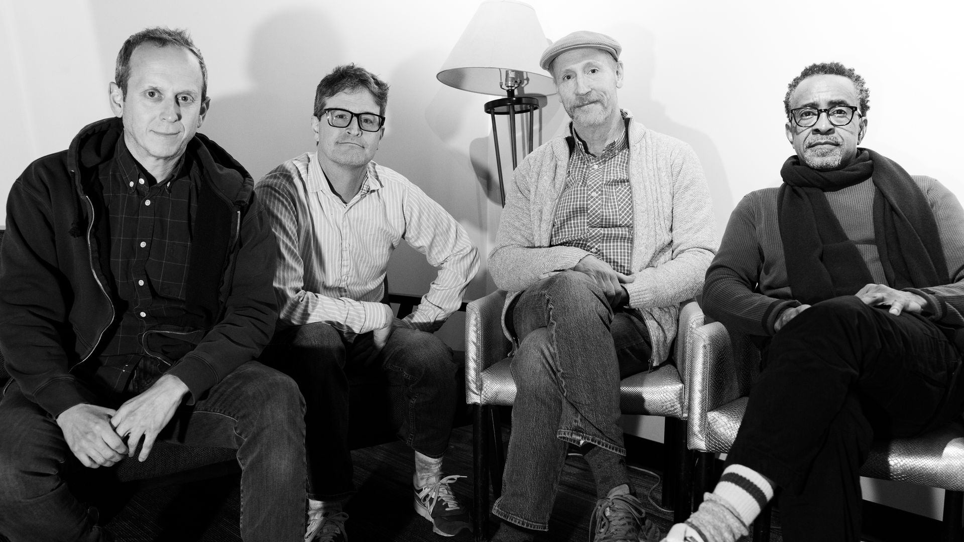 Four men sit in a room on chairs, posing for a photo. Left to right: dark-jacketed man, glasses in a striped shirt, cap-wearing man, and a scarf-wearing man. A floor lamp behind them.