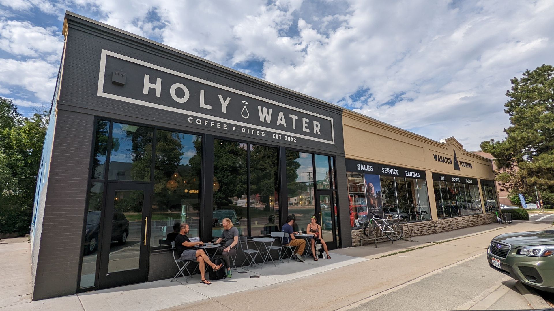 The storefront for Holy Water, a gray coffee shop, with people sitting at small round tables on the sidewalk.
