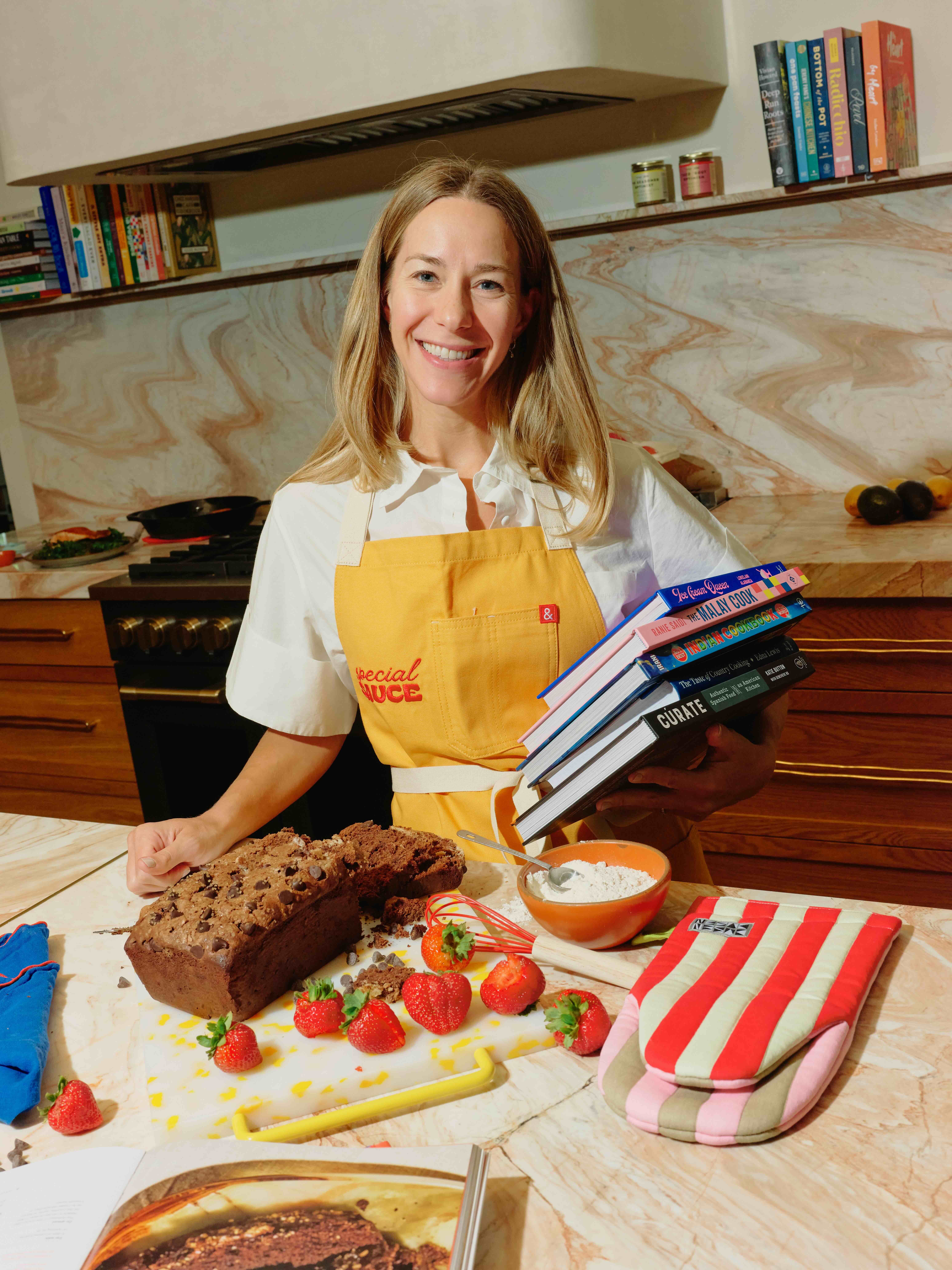 Woman in a white shirt holding cookbooks in front of a counter wearing a yellow apron that says "Special Sauce." 