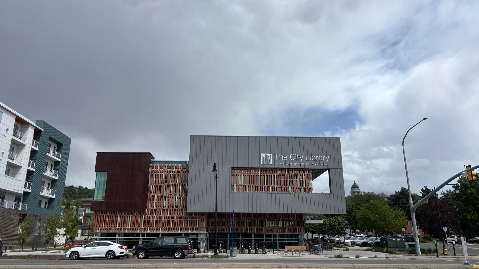 Urban street view of "The City Library" gray façade with orange lattice, a curved street lamp, and parked cars. Left shows white apartment blocks; stormy left sky and blue right.