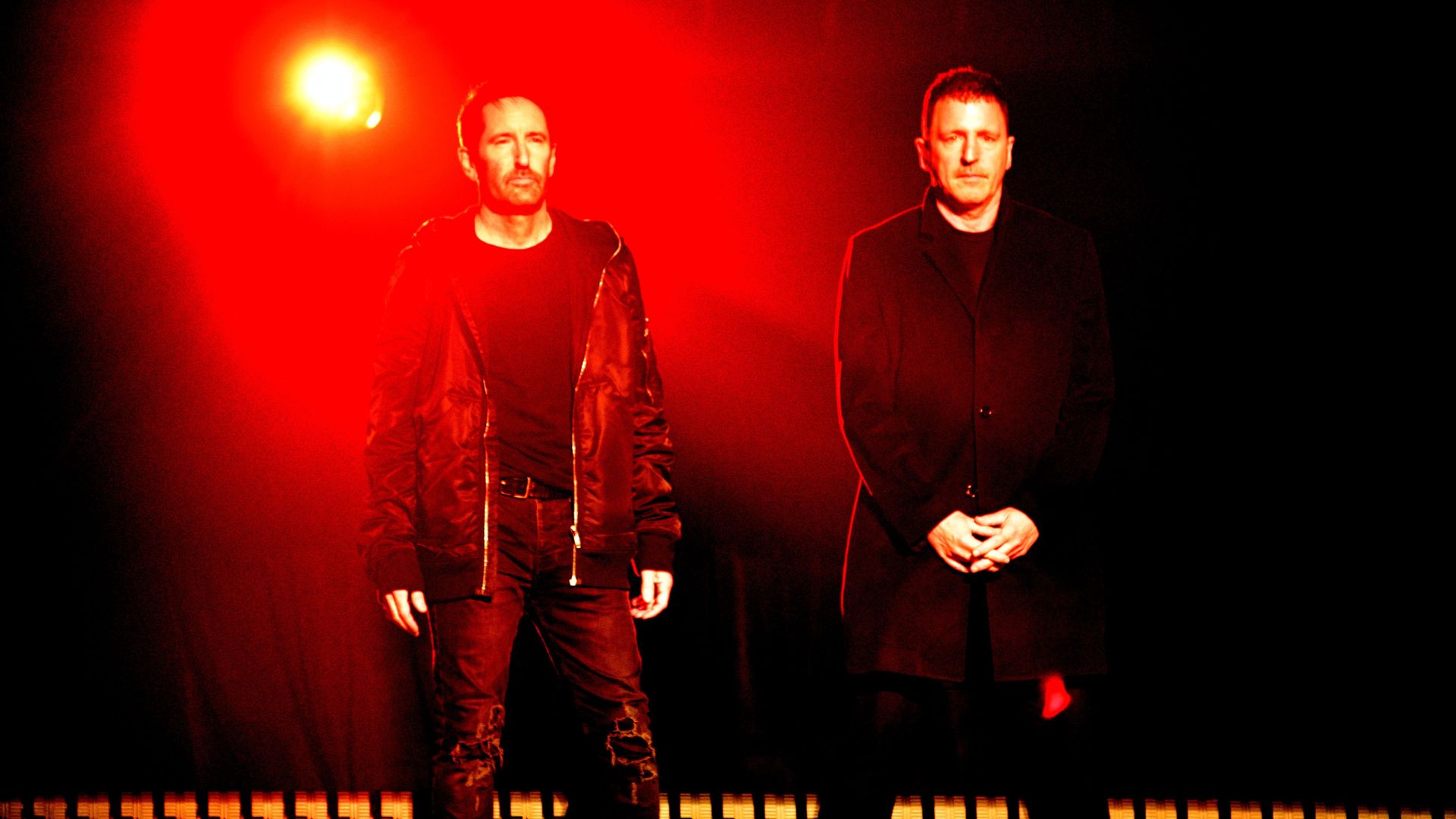 Musicians Trent Reznor and Atticus Ross stand on a stage lit by a red background.