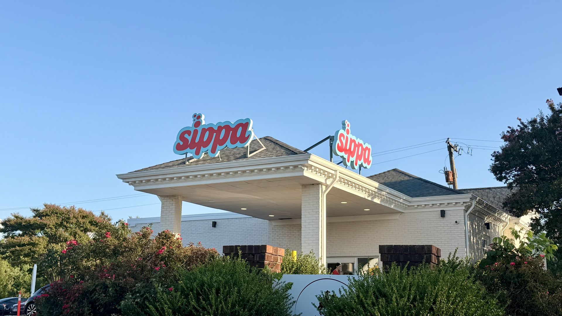 Clear blue sky over a white brick building with a covered entrance; two teal-outlined red "sippa" signs atop the roof, and greenery in the foreground.