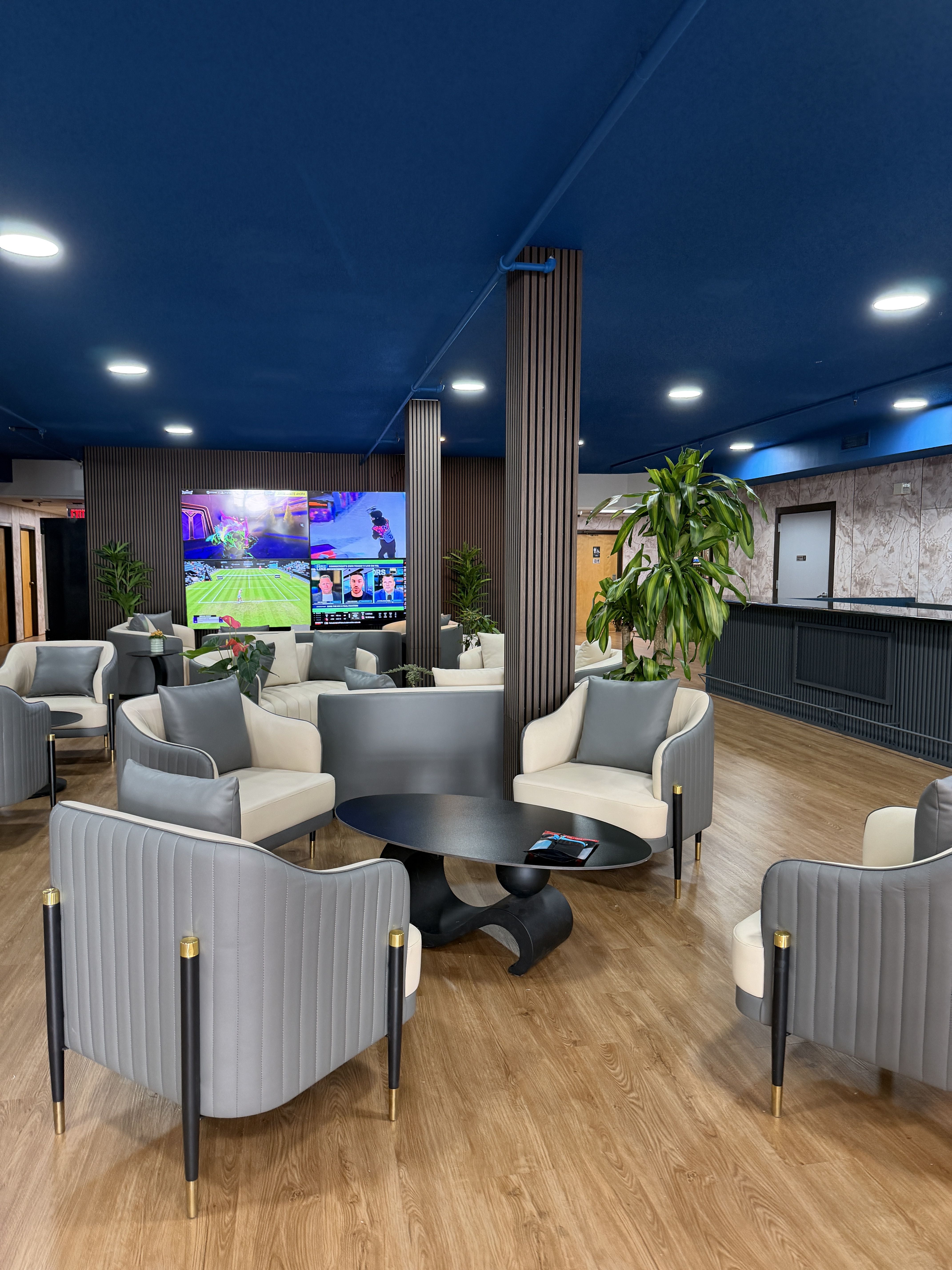 Modern lounge with a deep blue ceiling, round black coffee tables, gray and cream armchairs, tall wooden slat columns, a wall of screens showing sports, and several potted plants.