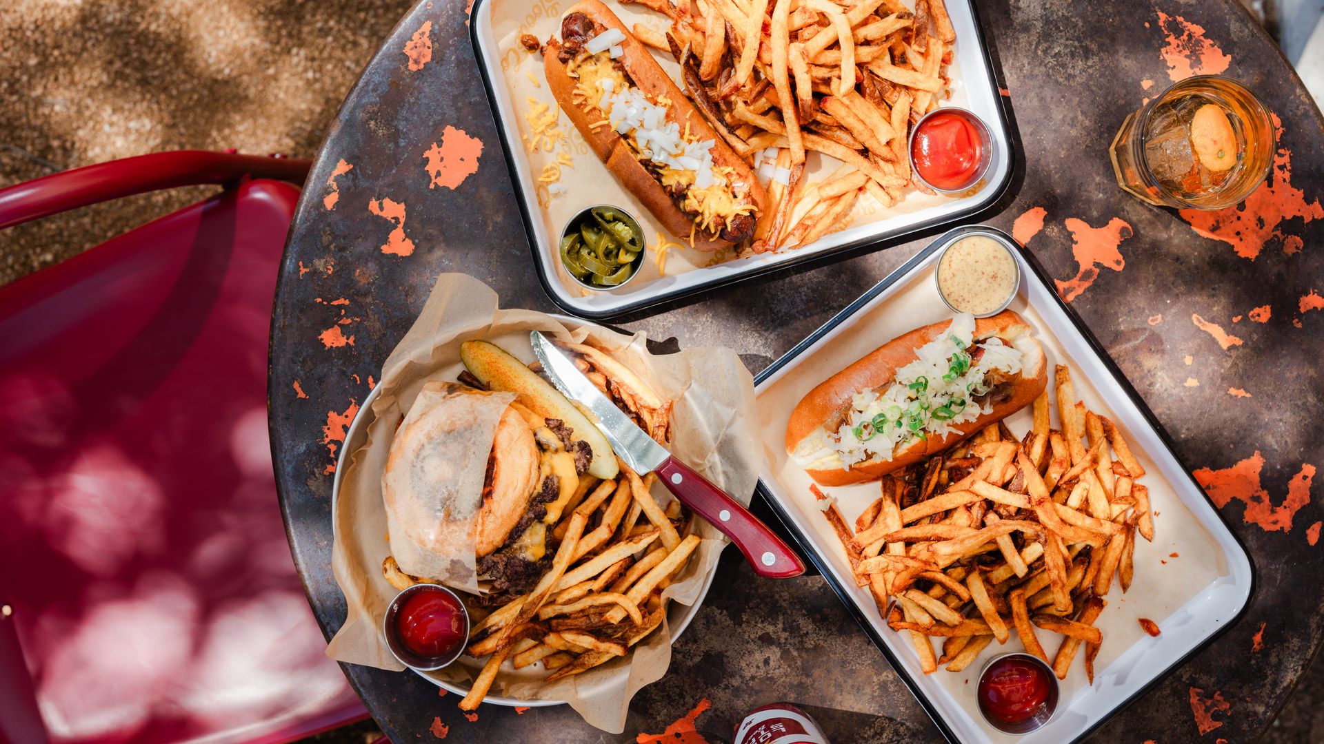 A view from above of a rusty table filled with three plates: Two with hot dogs with various fixings and fries, and one with a burger and fries. A cocktail glass sits on the side.