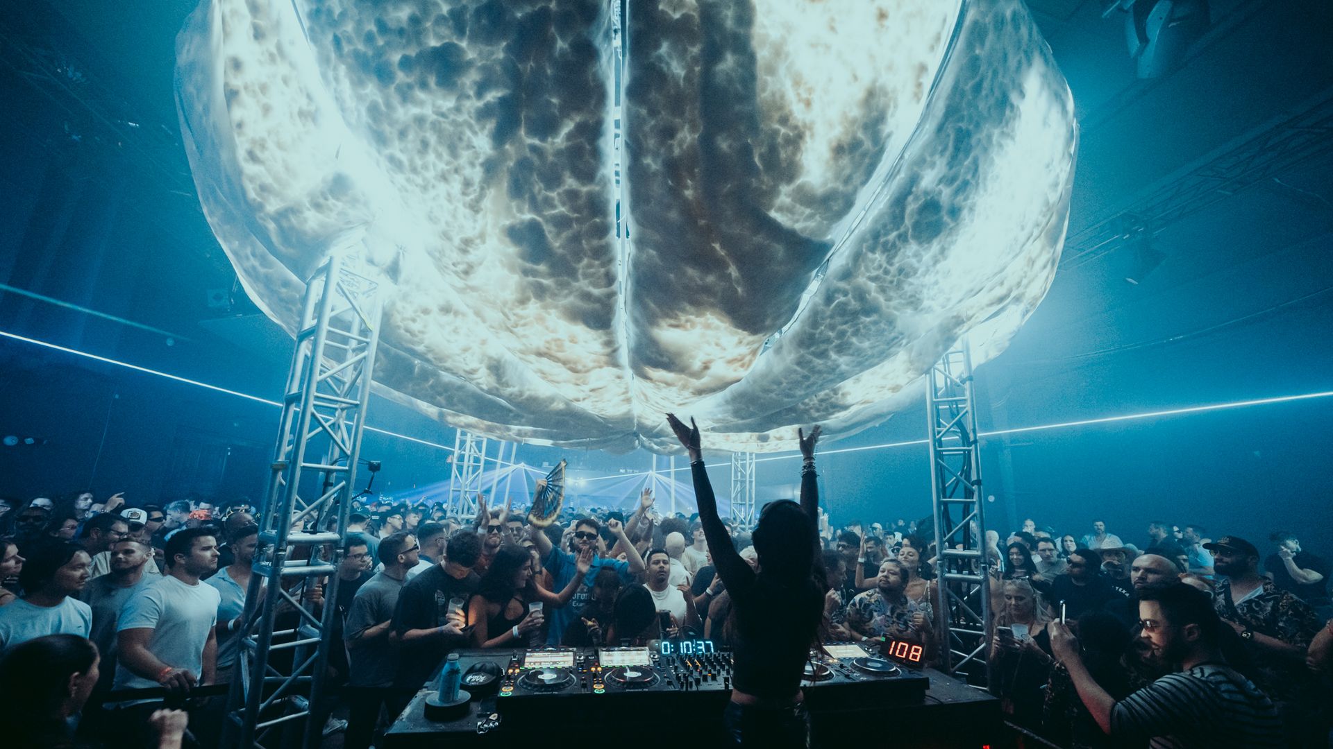 DJ performs at a nightclub with a colossal inflatable cloud overhead, blue lighting, and a packed crowd raising hands while the DJ booth glows with equipment.