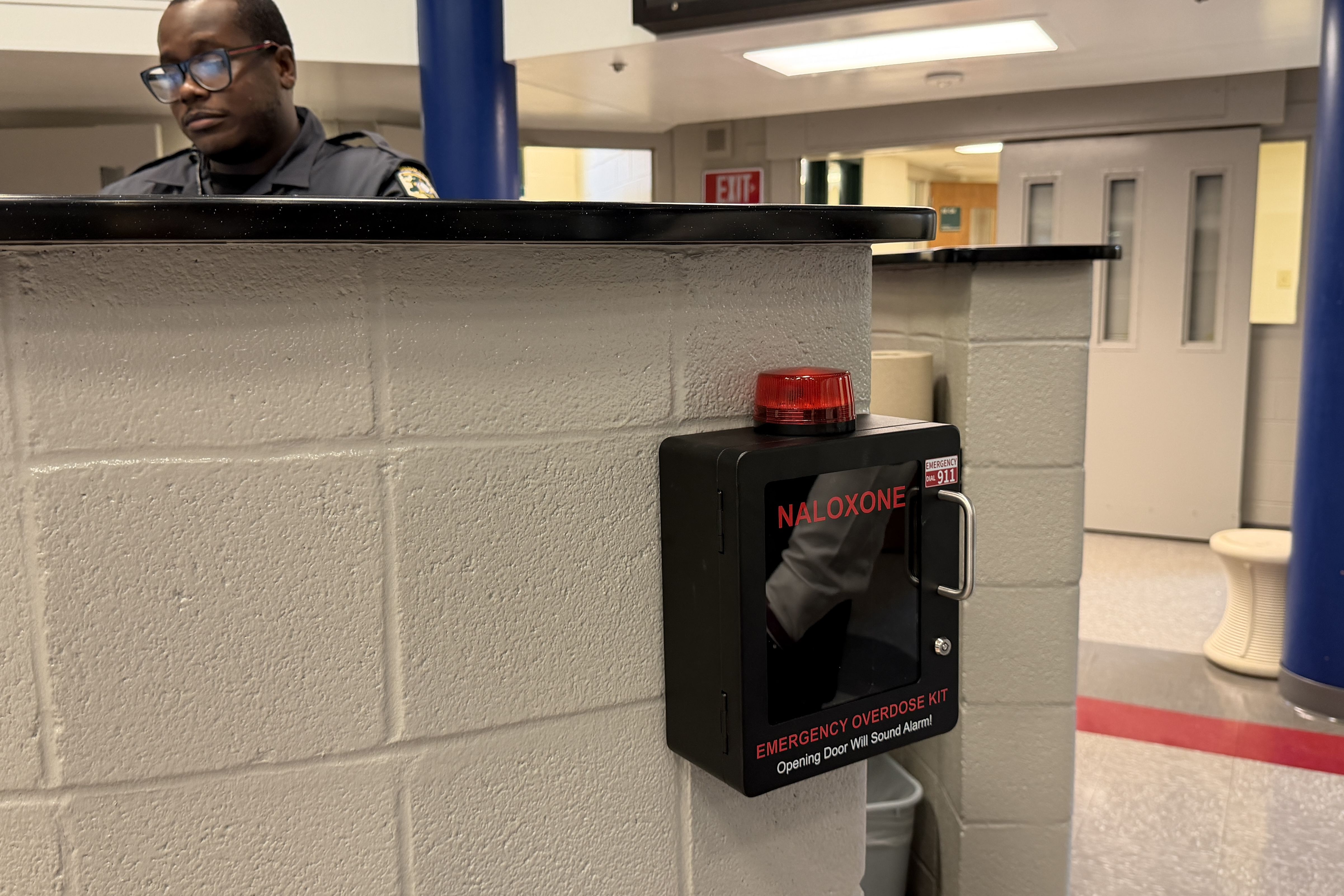 A security guard wearing glasses stands behind a gray counter in a building interior; a red Naloxone kit sits on the counter, with a blue pillar and an exit sign visible.