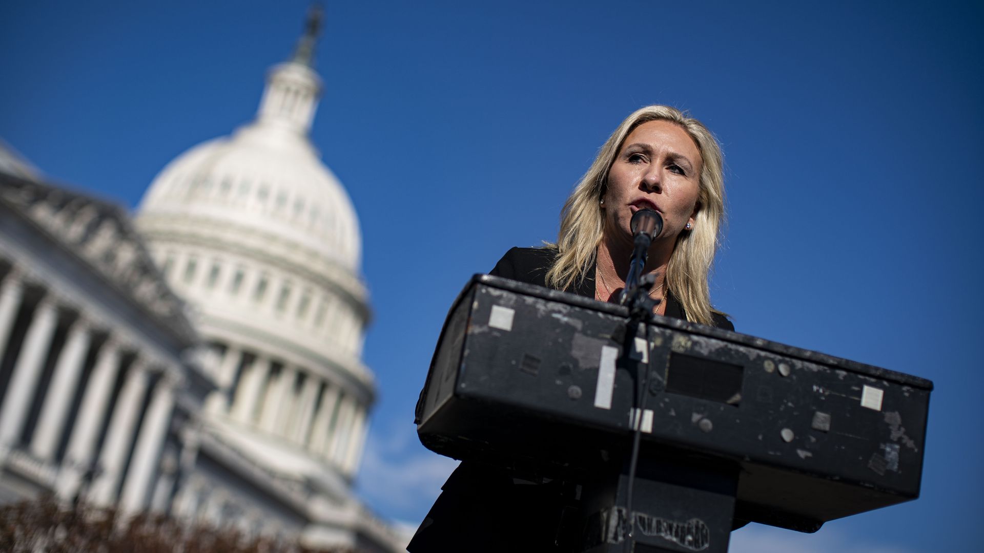 Picture of Marjorie Taylor Greene speaking behind a podium with Capitol Hill in the background