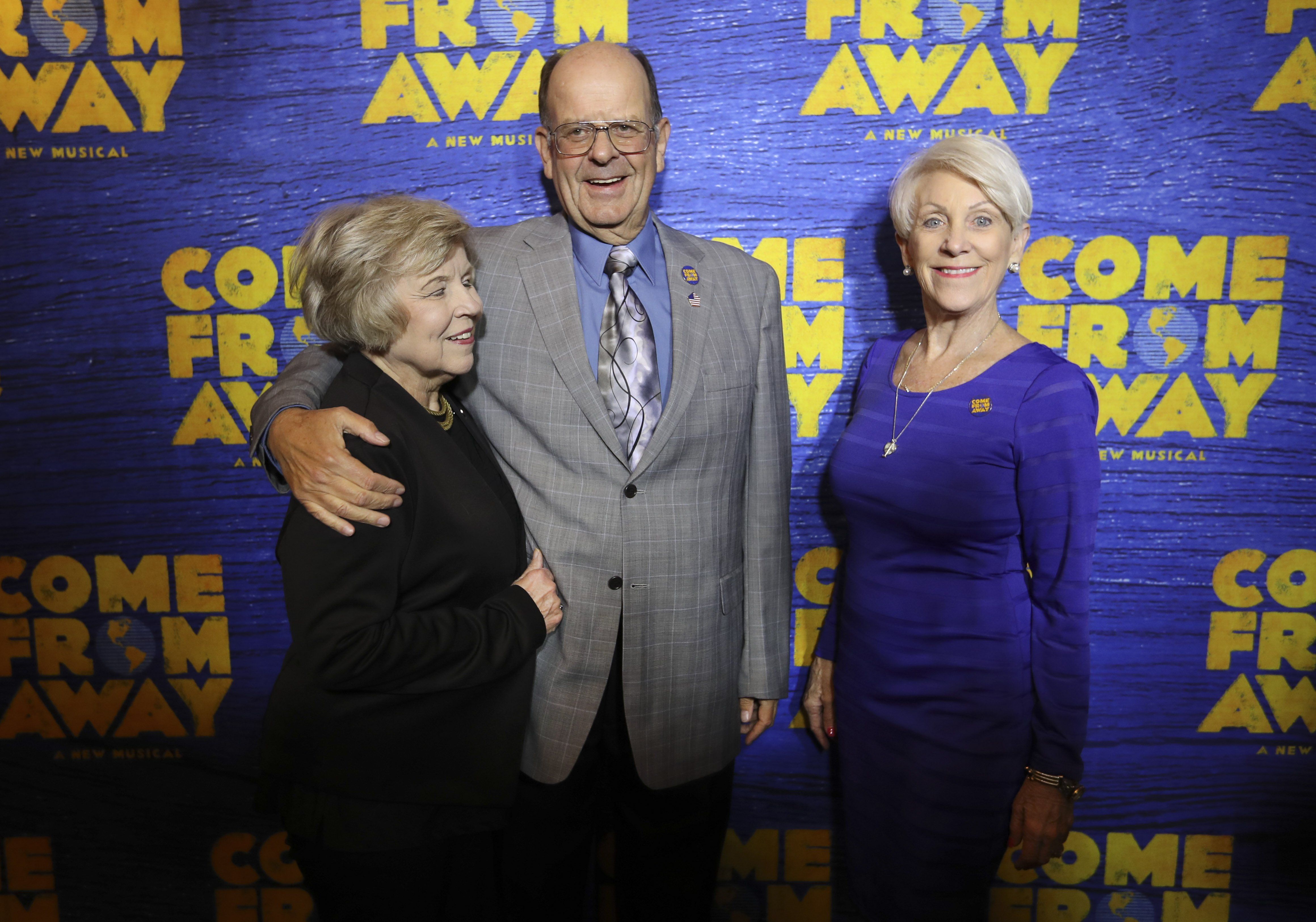 Three people pose with a blue backdrop that says "COME FROM AWAY" in bold yellow. Center: a man in a gray suit; flanked by two women in dark and purple outfits.