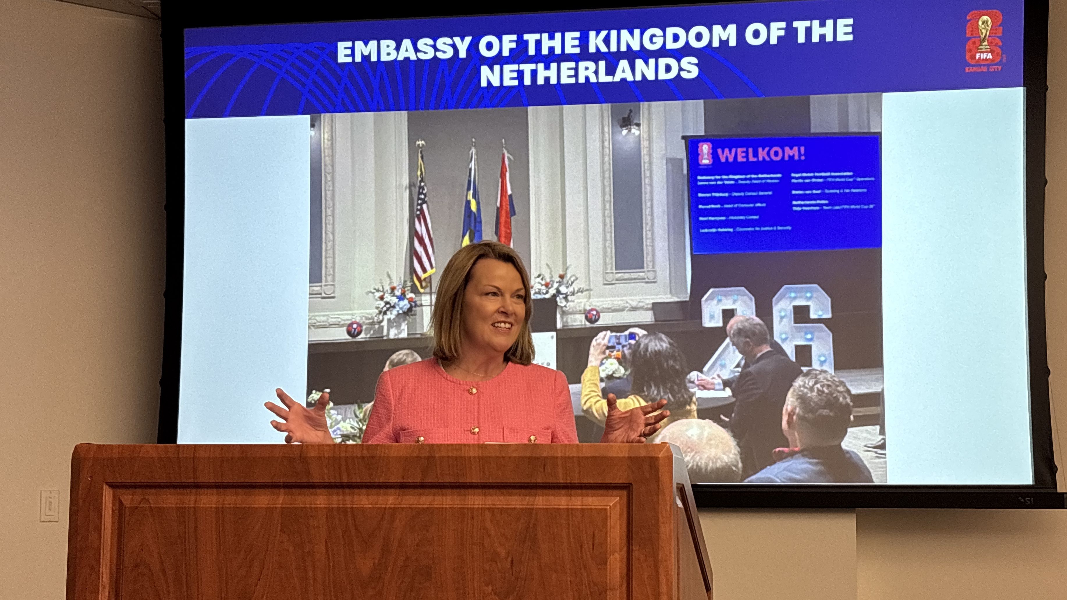 A woman in a pink jacket speaks at a wooden podium with a screen behind her reading "Embassy of the Kingdom of the Netherlands" and displaying flags and a welcome message.