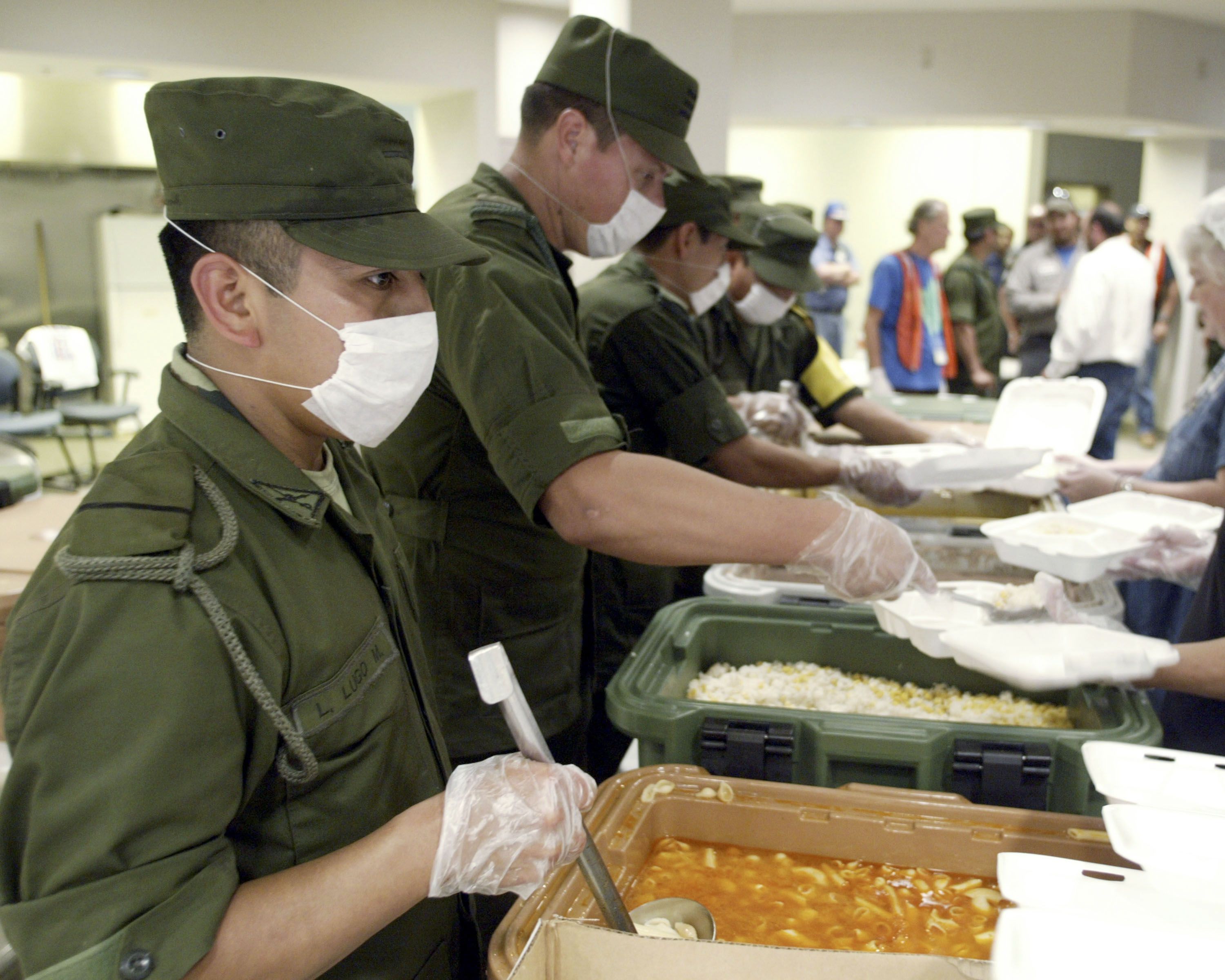 Men in green uniforms and white masks serving food from large containers into white takeout boxes in a busy indoor setting with people in the background.