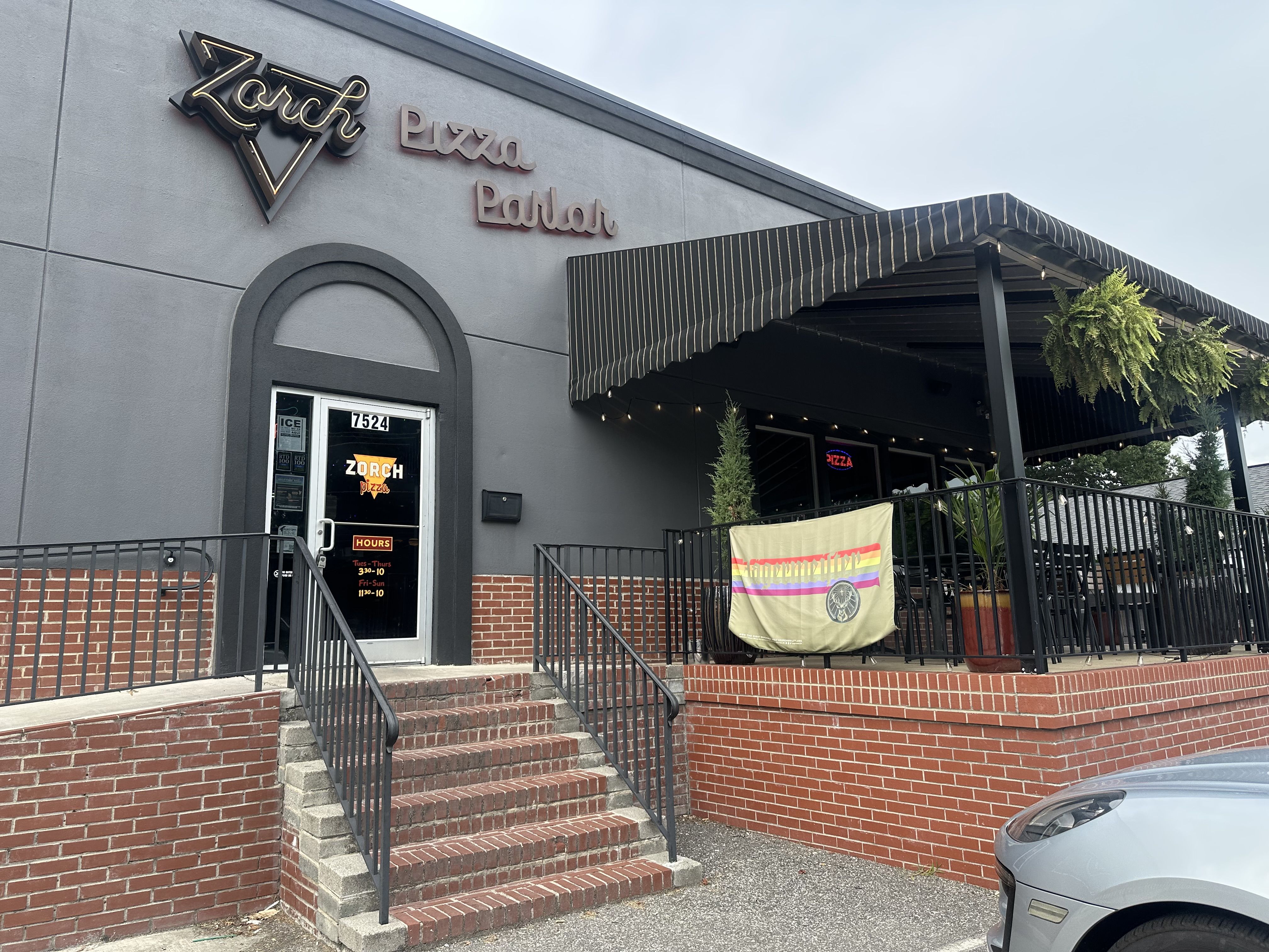 Gray, single-story building with a rounded arched doorway and a Zorch sign next to Pizza Parlor. A parking lot with red umbrellas, two cars, and a blue sky with trees.