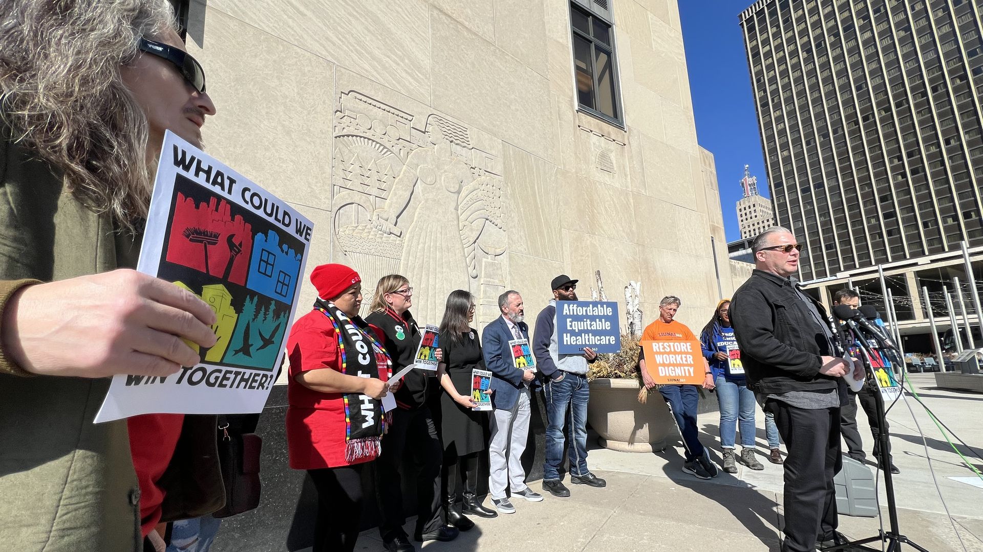 A man with light skin wearing sunglasses holds a colorful sign reading "What Could We Win Together?" as other people wearing bright-colored clothing stand in front of a stone wall for a press conference.