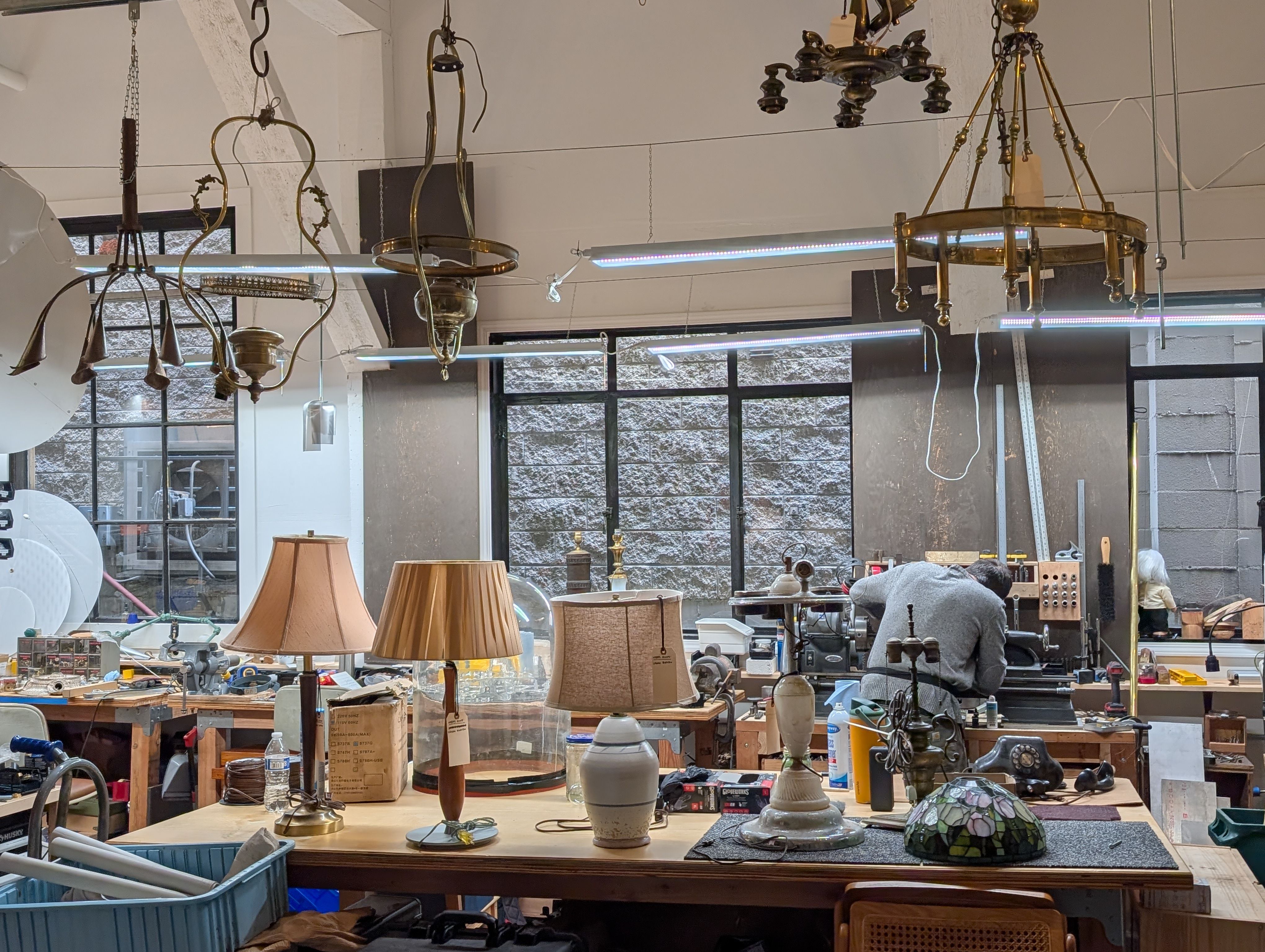 Vintage lighting shop: brass chandeliers hang from the ceiling alongside fluorescent tubes; cluttered tables hold lamps, glass displays, and tools, with a person in a gray hoodie near the back.