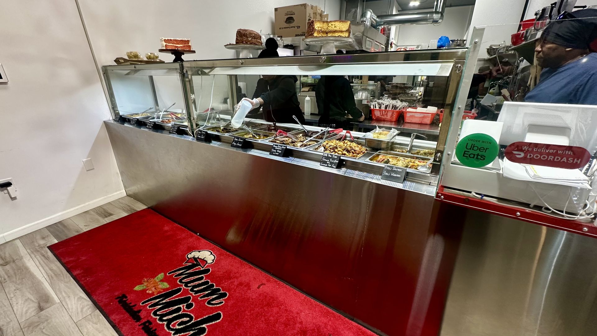 The interior of Mum Mich Trinidadian restaurant in Dorchester, where one worker adds sauce to a tray of food behind the ordering counter and another handles the register.