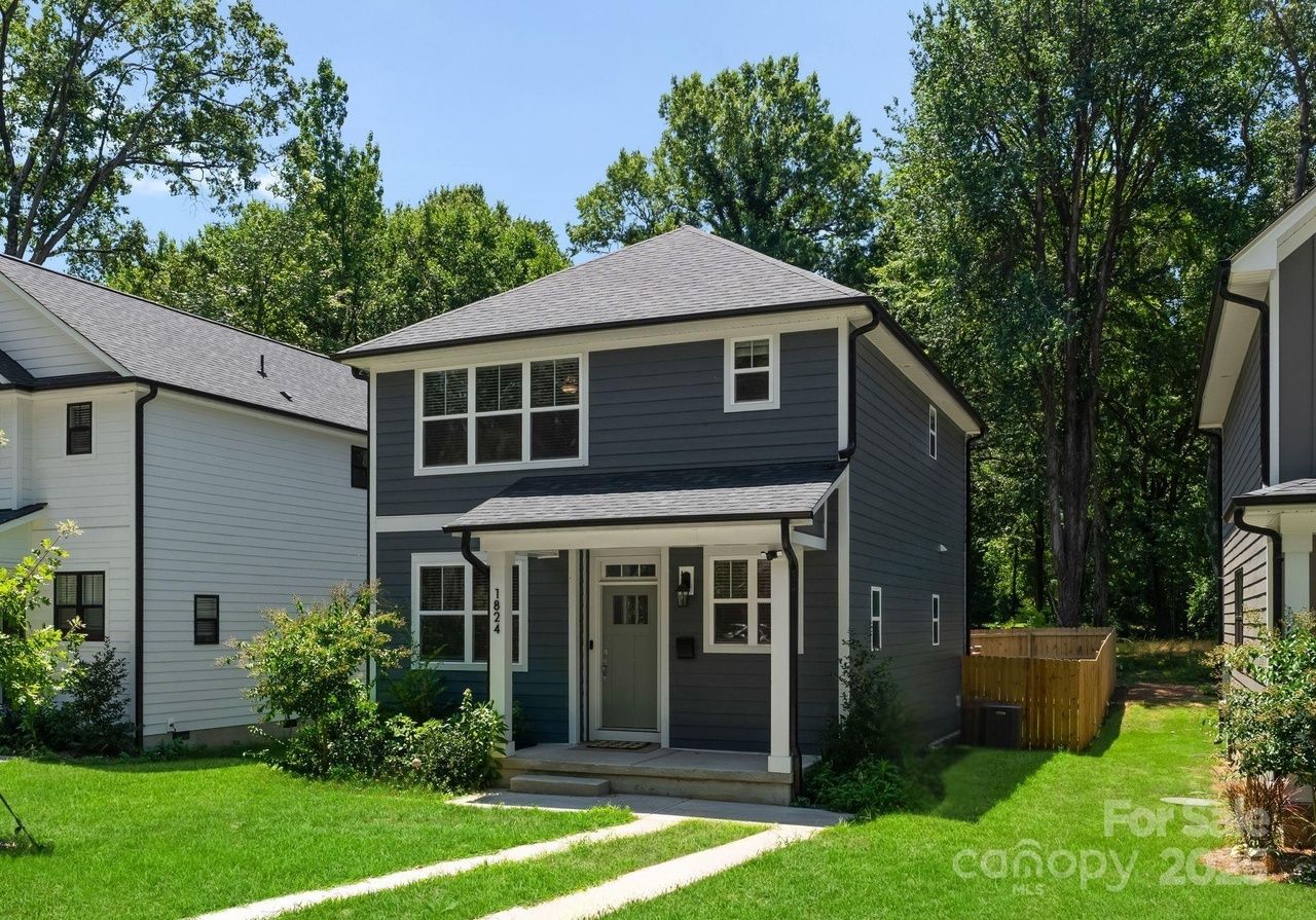 Two-story gray house with white trim, porch, and large windows, surrounded by green grass and trees under a clear blue sky. Adjacent white houses visible on both sides.