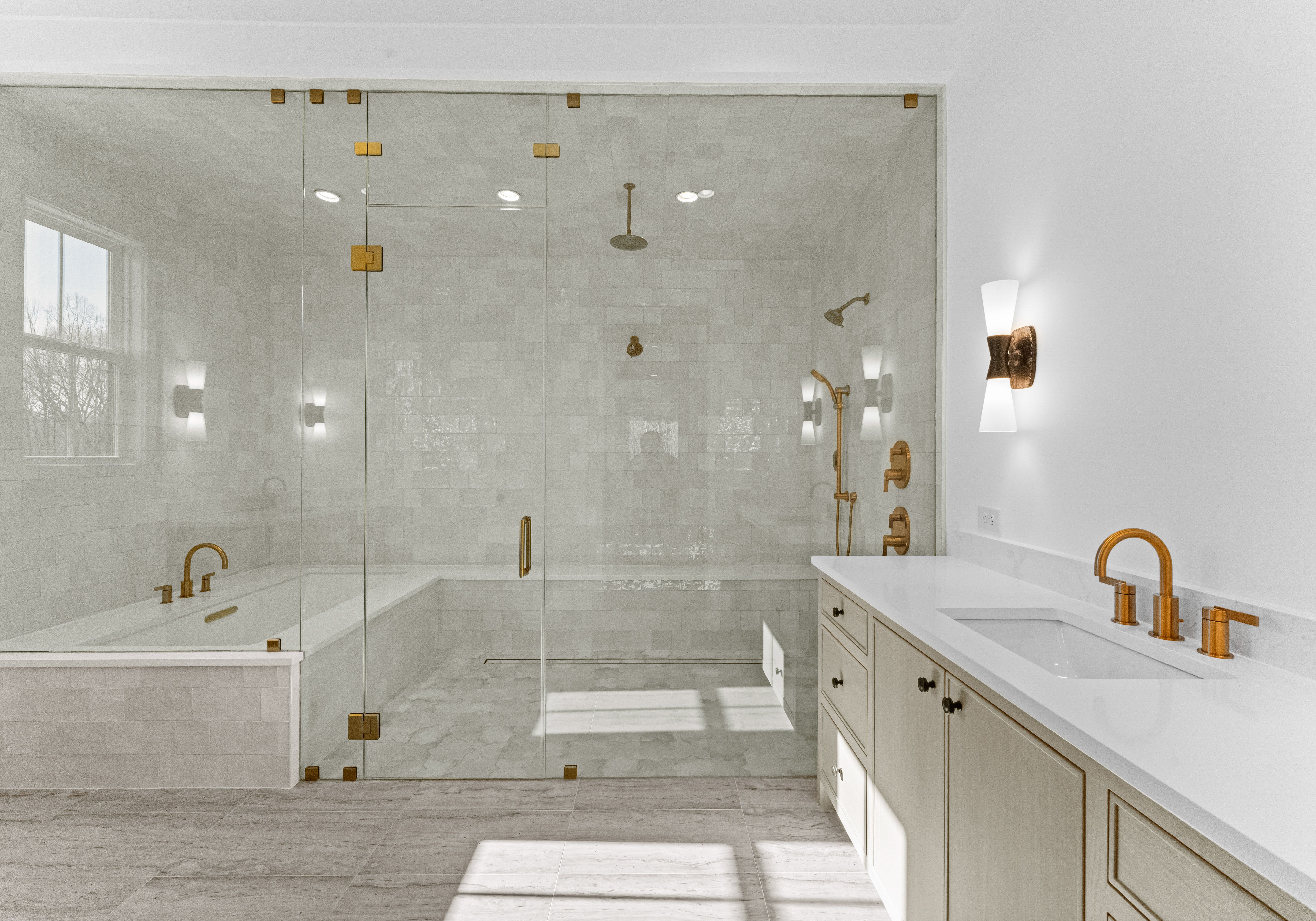 Bright modern bathroom with large glass shower enclosure featuring gold fixtures, white tiled walls, a built-in tub, and a white vanity with gold faucet and wall sconces.